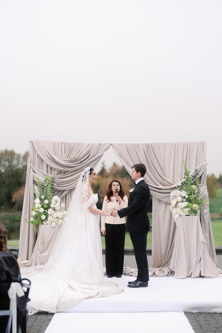 Outdoor wedding ceremony with a couple exchanging vows under a draped arch adorned with white flowers, officiated by a woman holding a microphone.