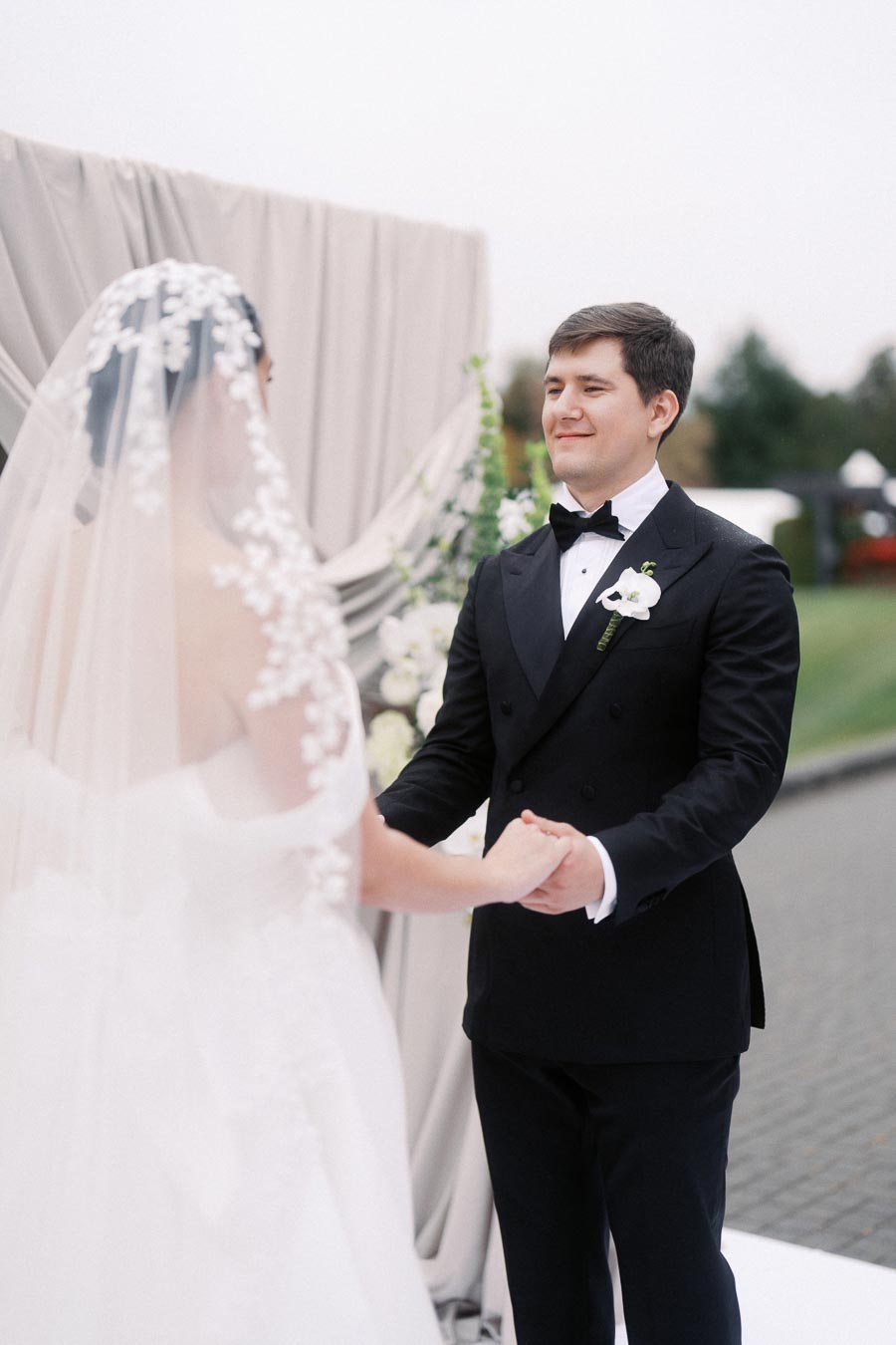 Bride and groom holding hands during an outdoor wedding ceremony, with the groom in a black tuxedo smiling at the bride, who is wearing a lace-embellished veil.