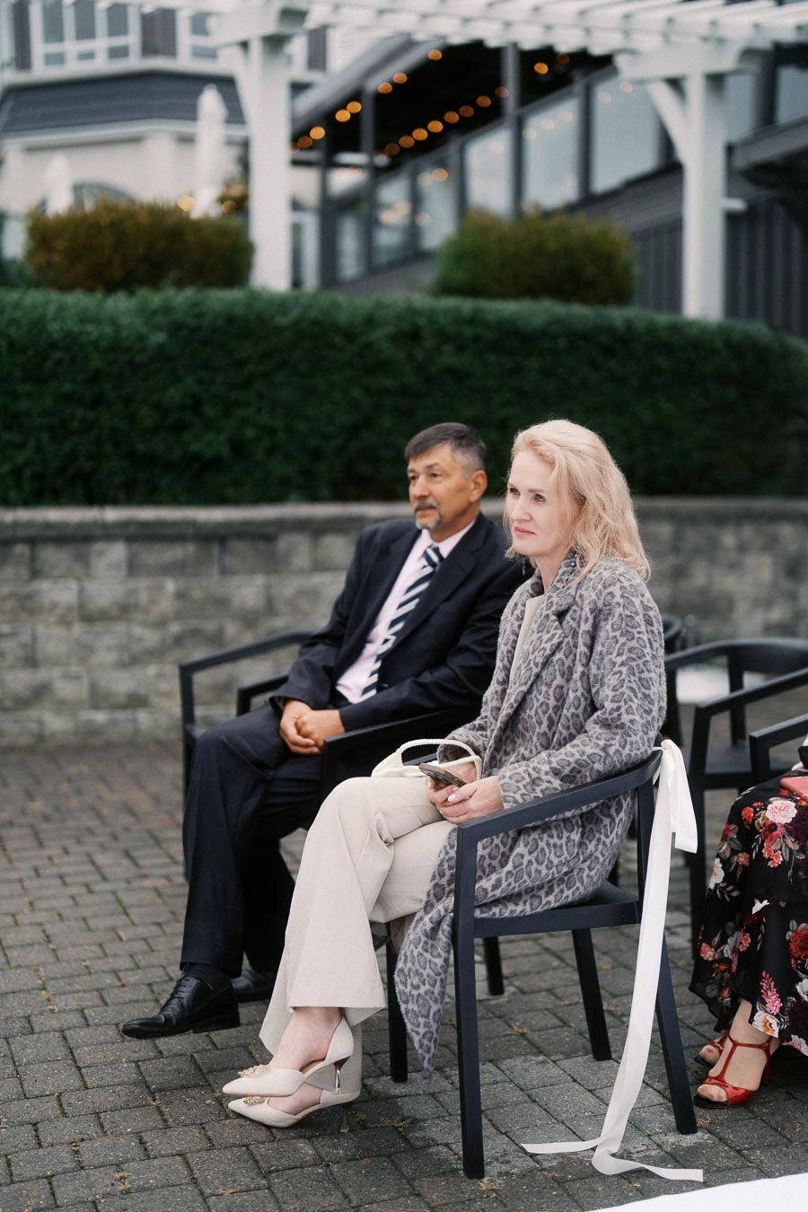 Two people seated outdoors during an event, dressed in formal attire, with greenery and a modern building in the background.