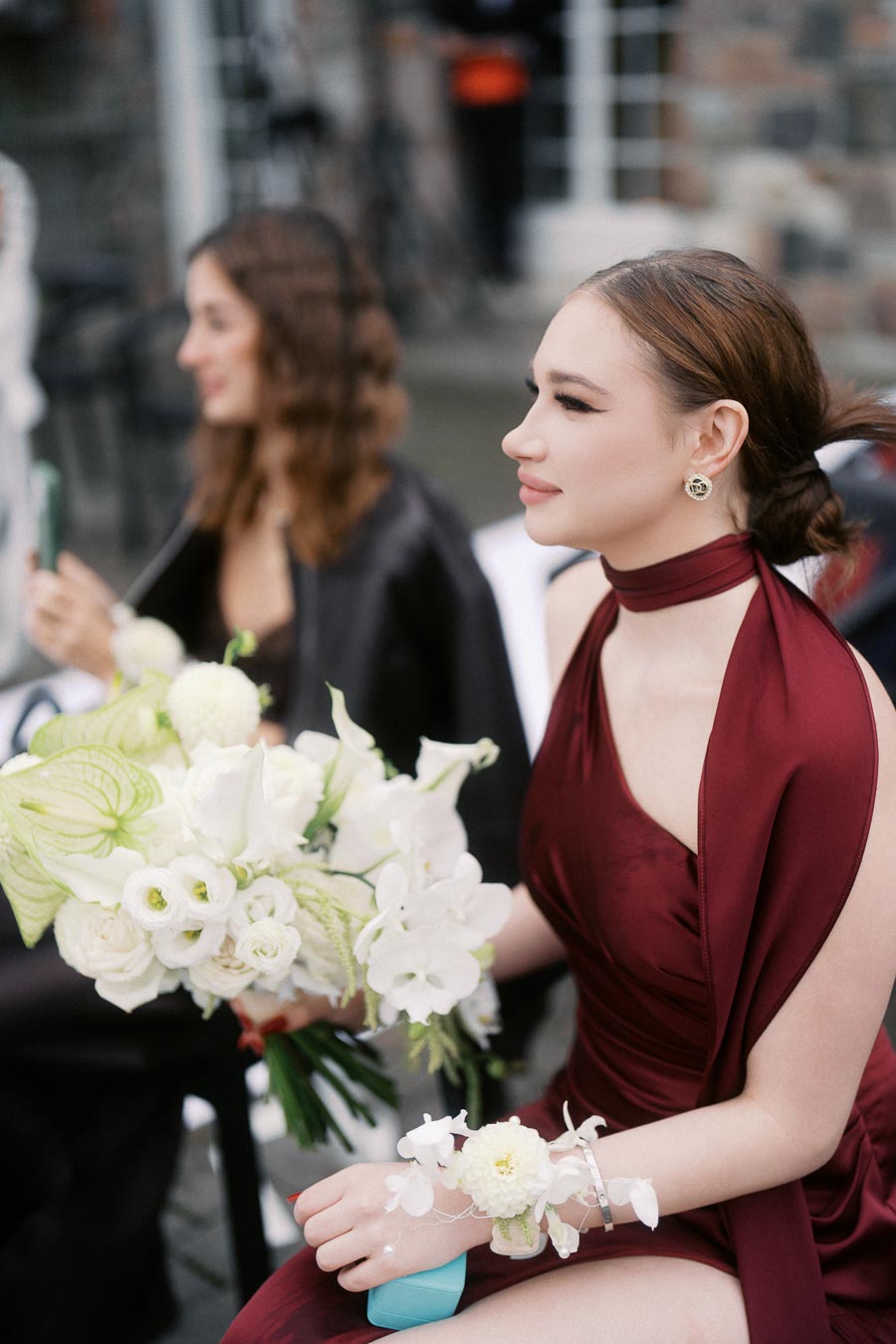 Elegant woman in a burgundy dress holding a bouquet of white flowers, sitting outdoors.