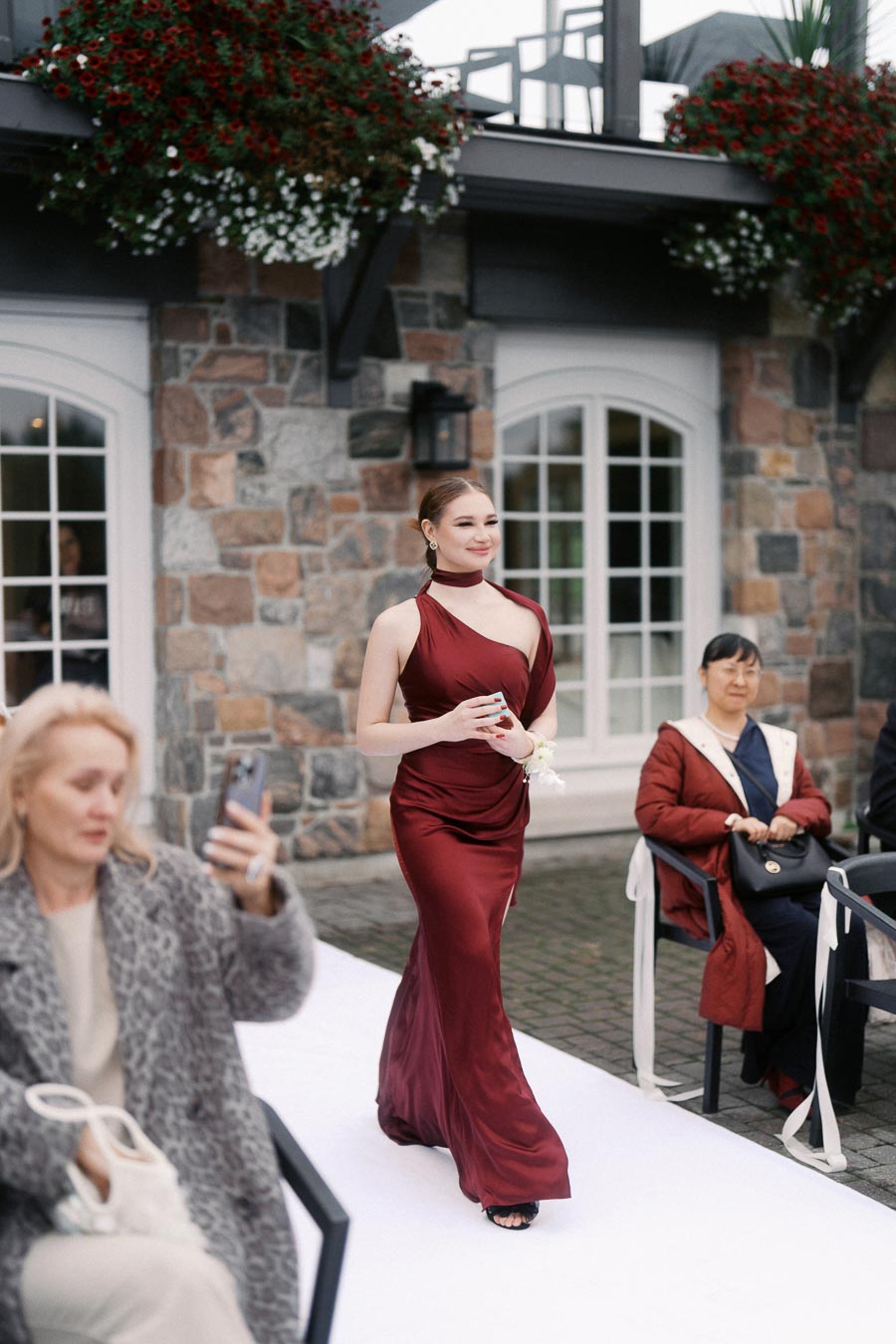 Elegant woman in a burgundy gown walking on a white runway outdoors, with onlookers seated nearby, against a stone building backdrop adorned with hanging flowers.