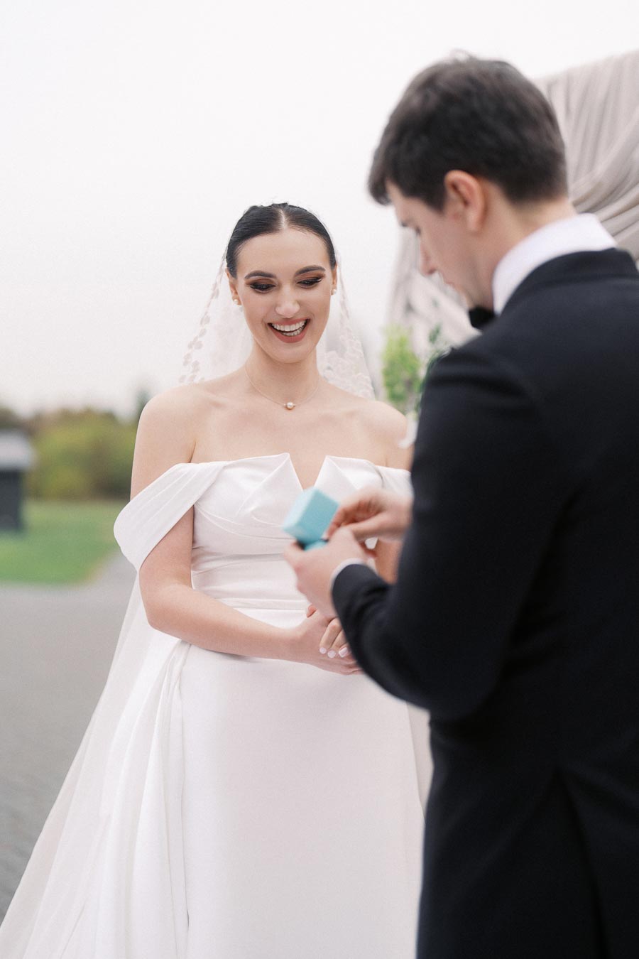 Bride in elegant white gown smiling as groom opens a small blue gift box during an outdoor wedding ceremony.