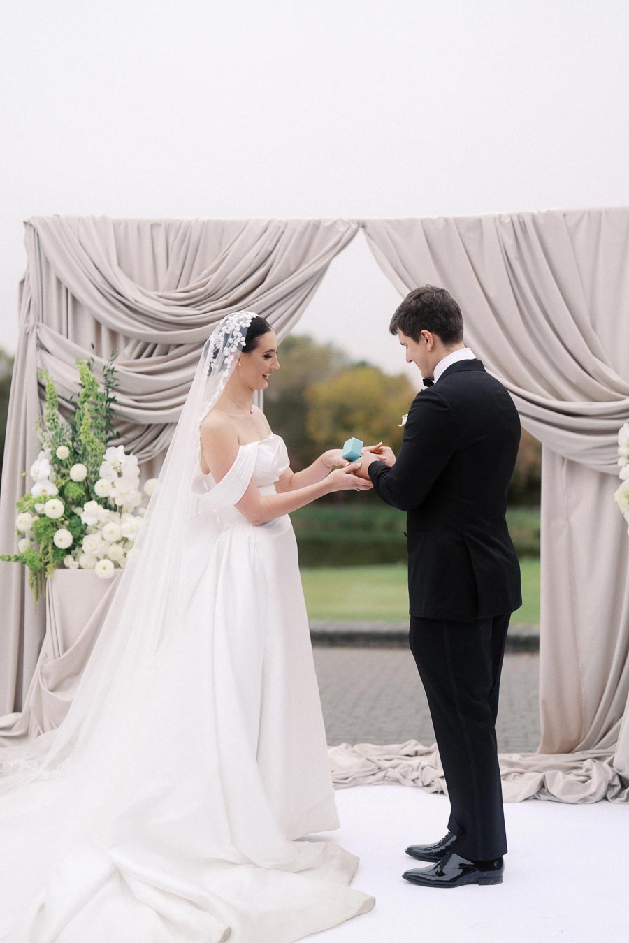 Bride and groom exchange vows in a beautiful outdoor ceremony, with elegant draped fabric and floral decorations in the background.