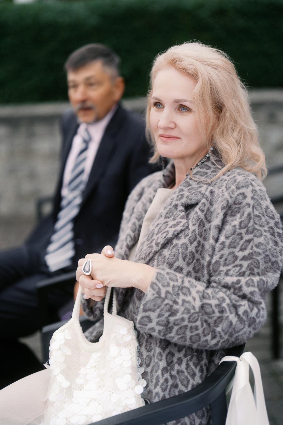 A woman with blonde hair wearing a leopard print coat holds a white sequined bag, sitting outdoors with a blurred man in the background.