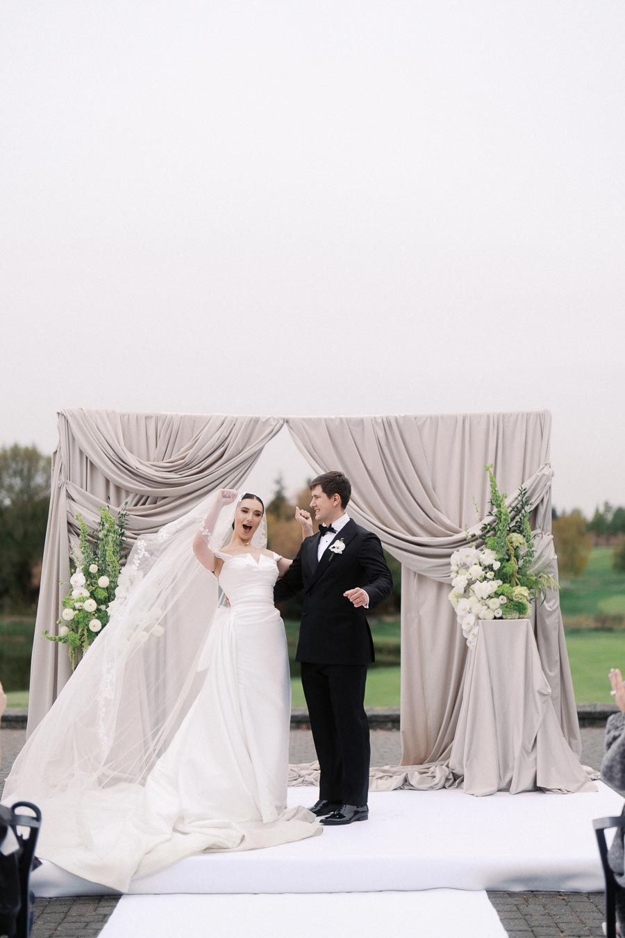 Bride and groom celebrating joyfully at outdoor wedding ceremony with elegant backdrop and floral arrangements.