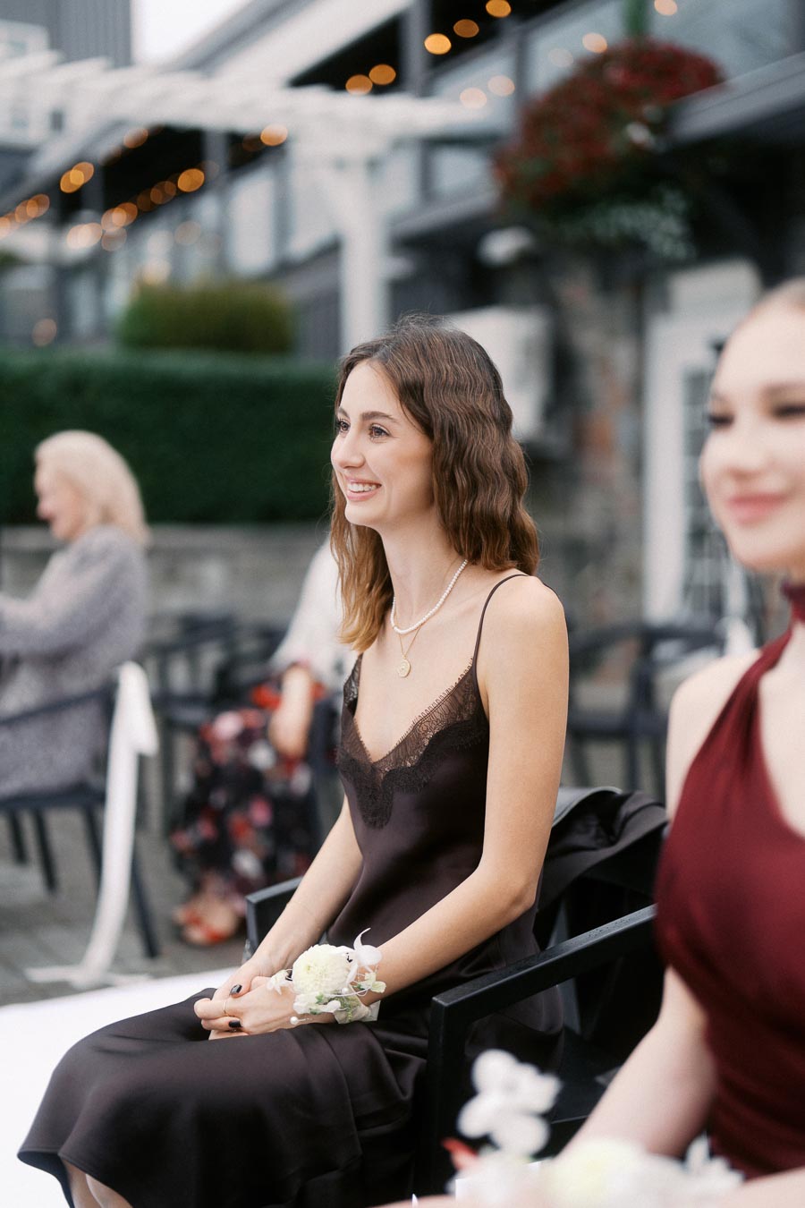 Young woman in a black dress sitting outdoors at an elegant event with blurred background.