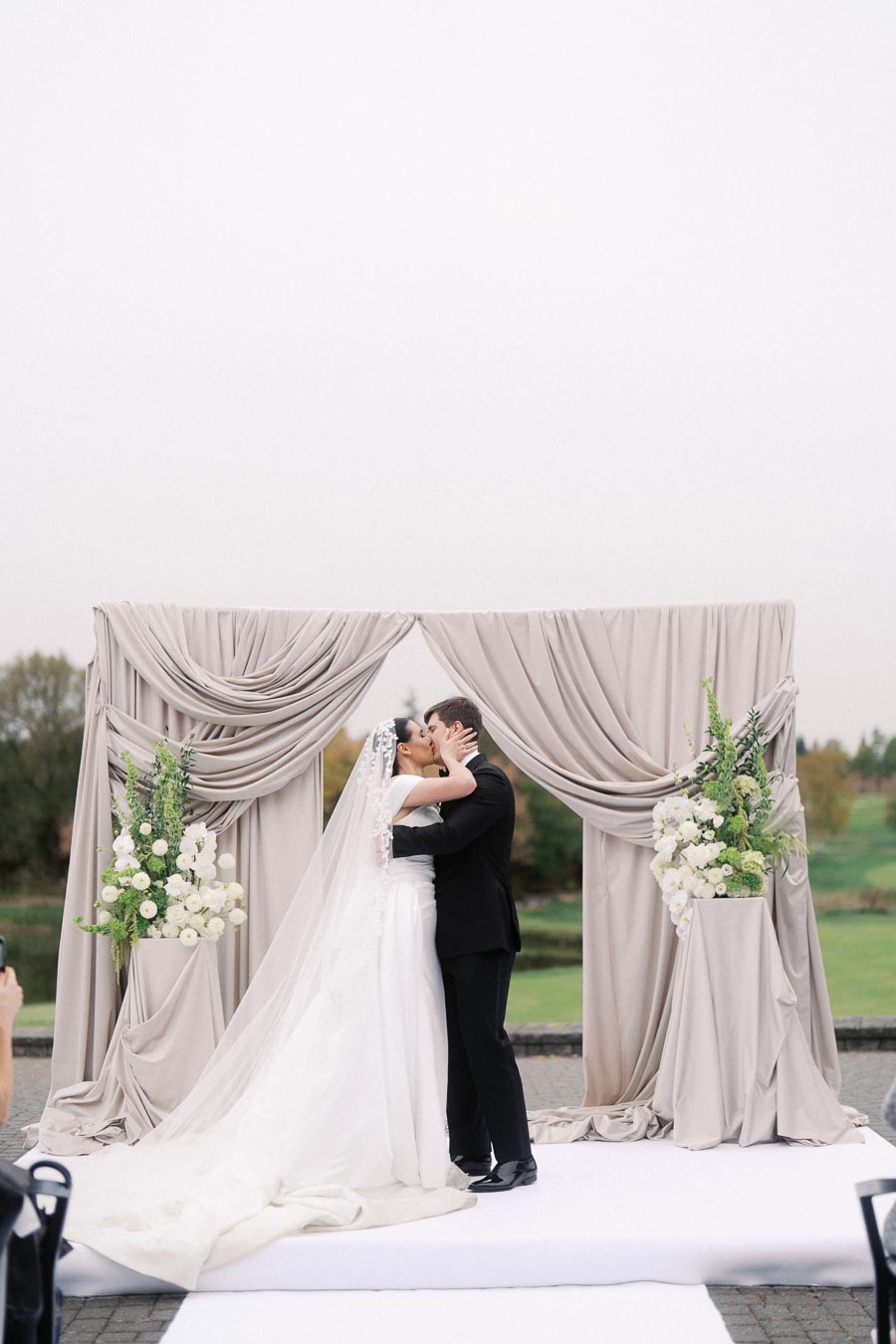 Bride and groom share a romantic kiss under an elegant draped arch with floral arrangements during their outdoor wedding ceremony.