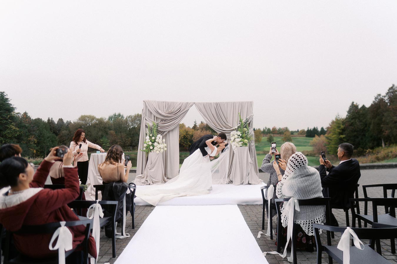 Outdoor wedding ceremony with bride and groom kissing under a decorated arch, surrounded by guests taking photos with smartphones.