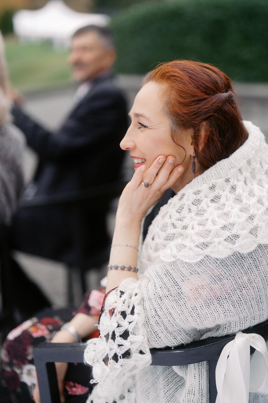 Woman in a white crocheted shawl smiling during an outdoor event, with a soft-focused person in the background and greenery surrounding the scene.
