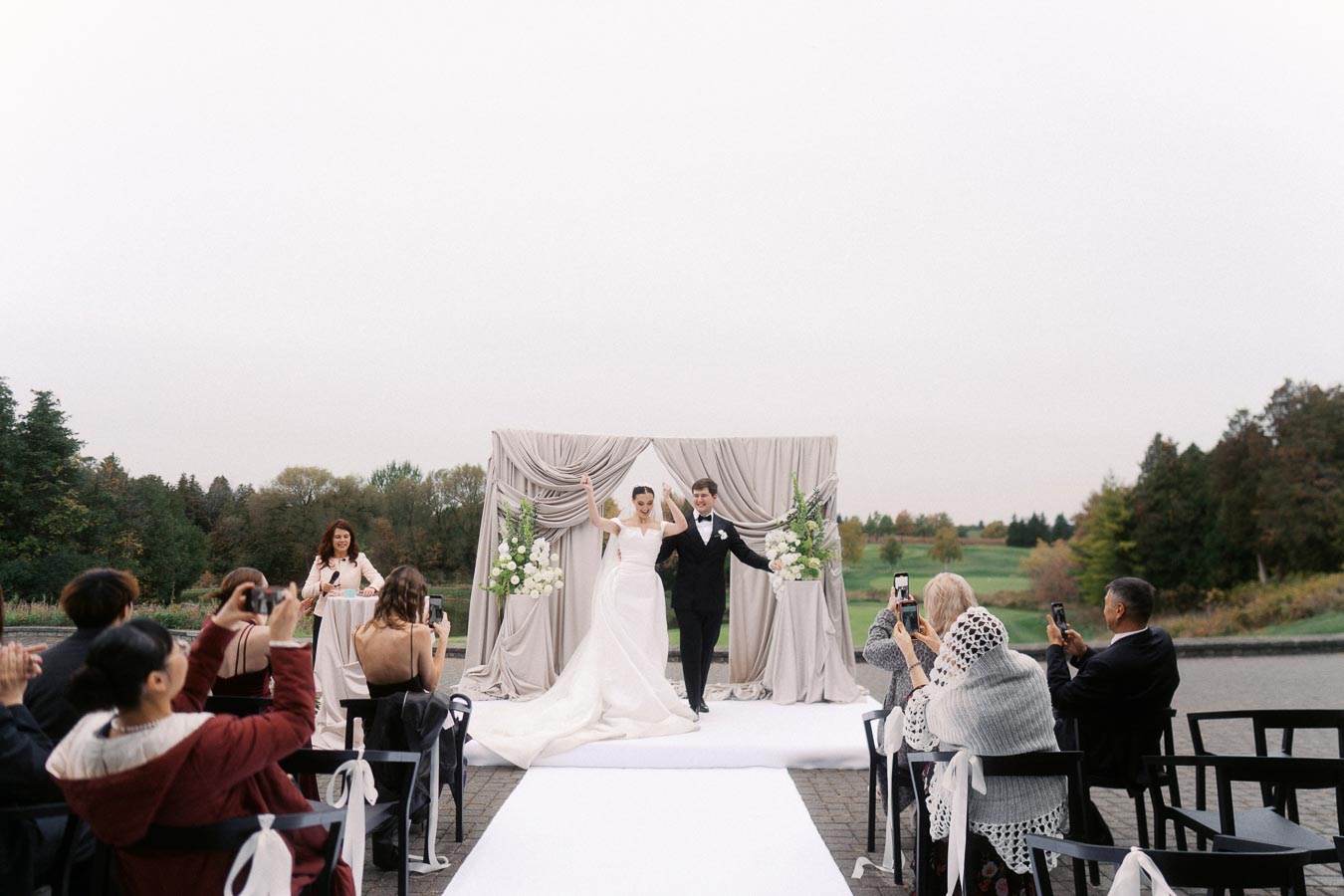 Outdoor wedding ceremony with a bride and groom walking down the aisle, surrounded by guests capturing the moment on their phones. The couple stands in front of an elegant backdrop with white floral arrangements, set against a picturesque landscape.