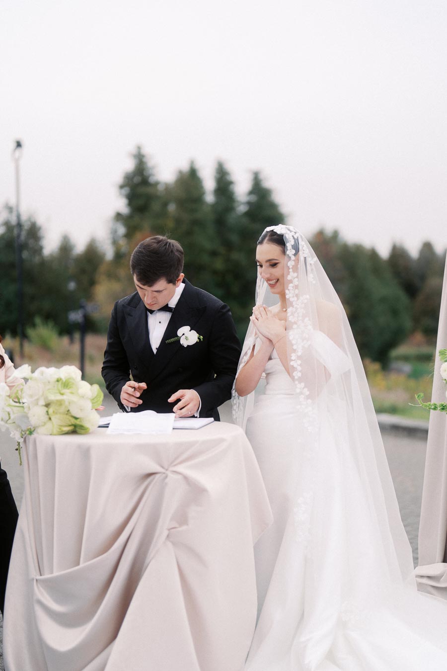Bride and groom signing wedding documents at outdoor ceremony table, surrounded by floral arrangements and greenery.