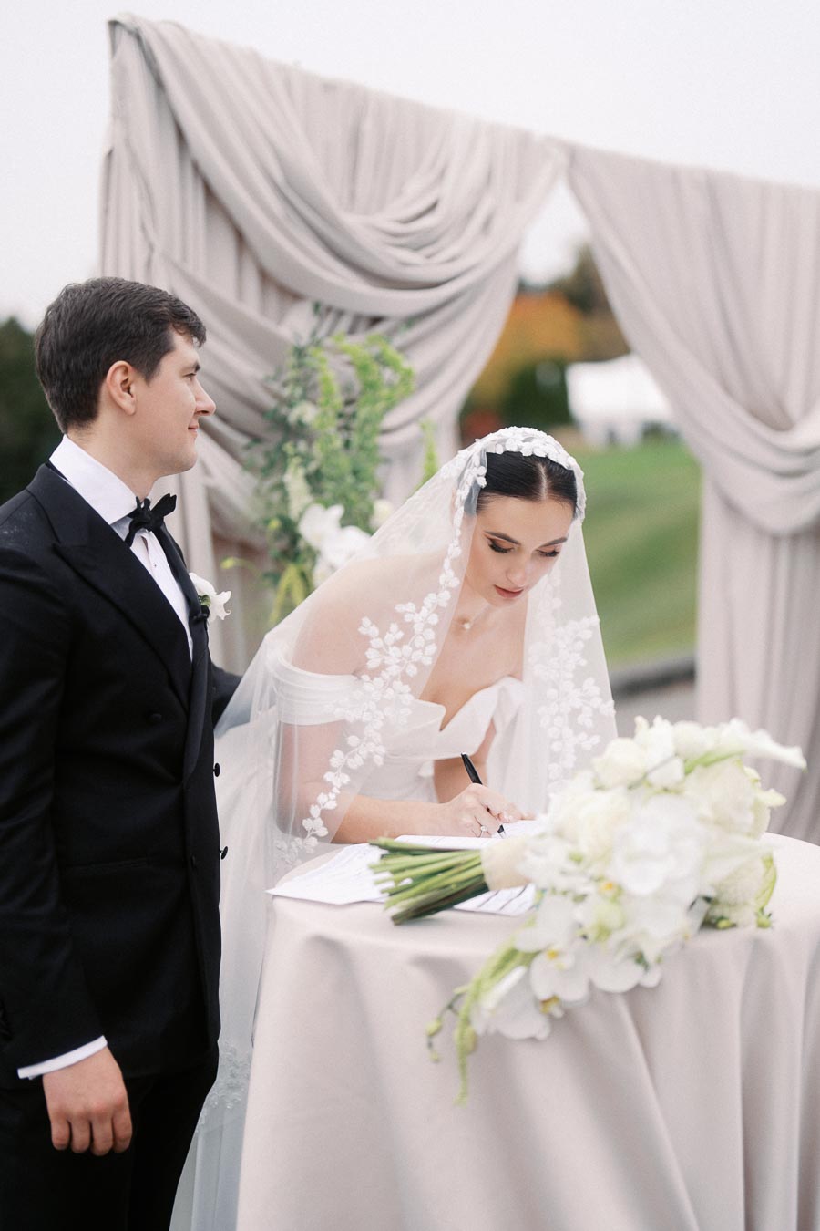 Bride signing a document at outdoor wedding ceremony, wearing a floral lace veil and off-shoulder gown, with groom in a black tuxedo standing beside her. Elegant floral arrangement in foreground, with draped backdrop and garden setting.