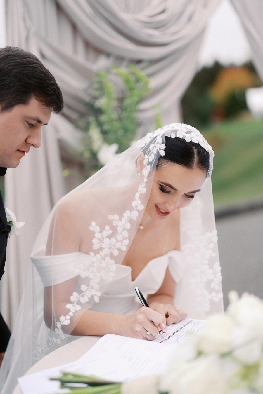 Bride signing marriage certificate at outdoor wedding ceremony, wearing a veil with floral embroidery, accompanied by groom, with elegant drapery and greenery in the background.