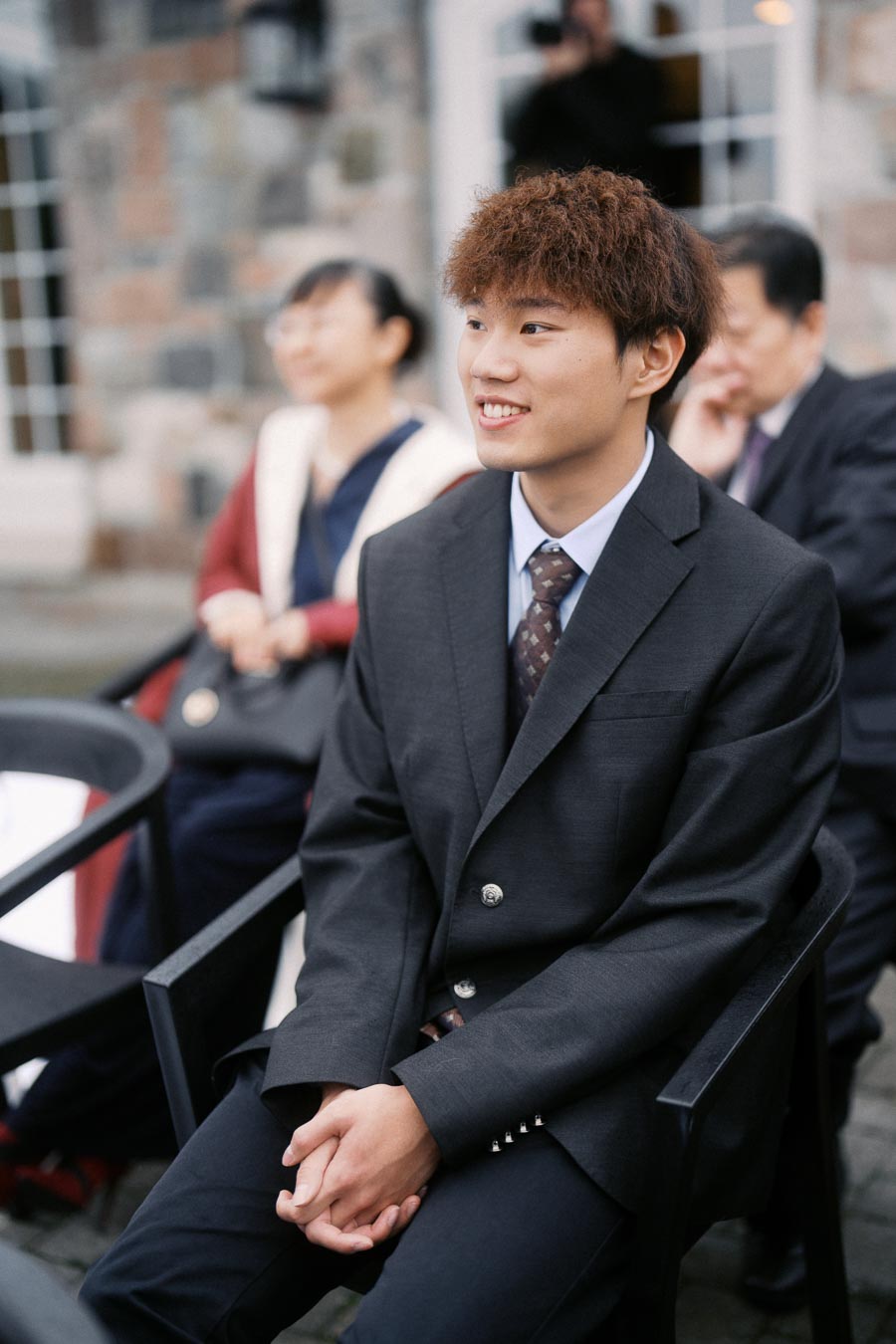 Young man in a suit seated outdoors at an event, smiling warmly, with other attendees blurred in the background.