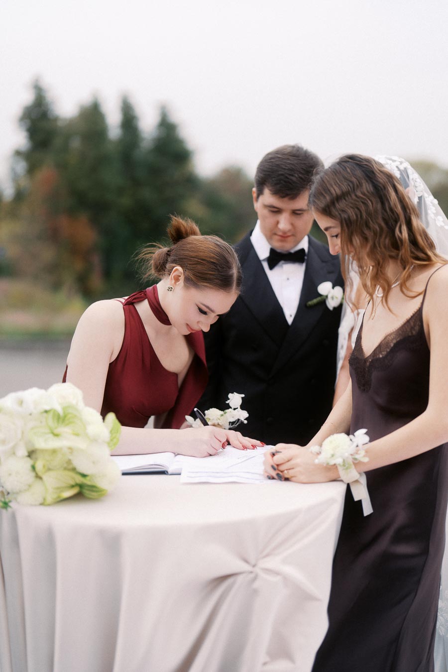Three elegantly dressed individuals, including a bridesmaid in a red dress, a groomsman in a tuxedo, and a woman in a dark dress with a corsage, are signing a marriage document on a white table adorned with a bouquet of flowers outdoors.