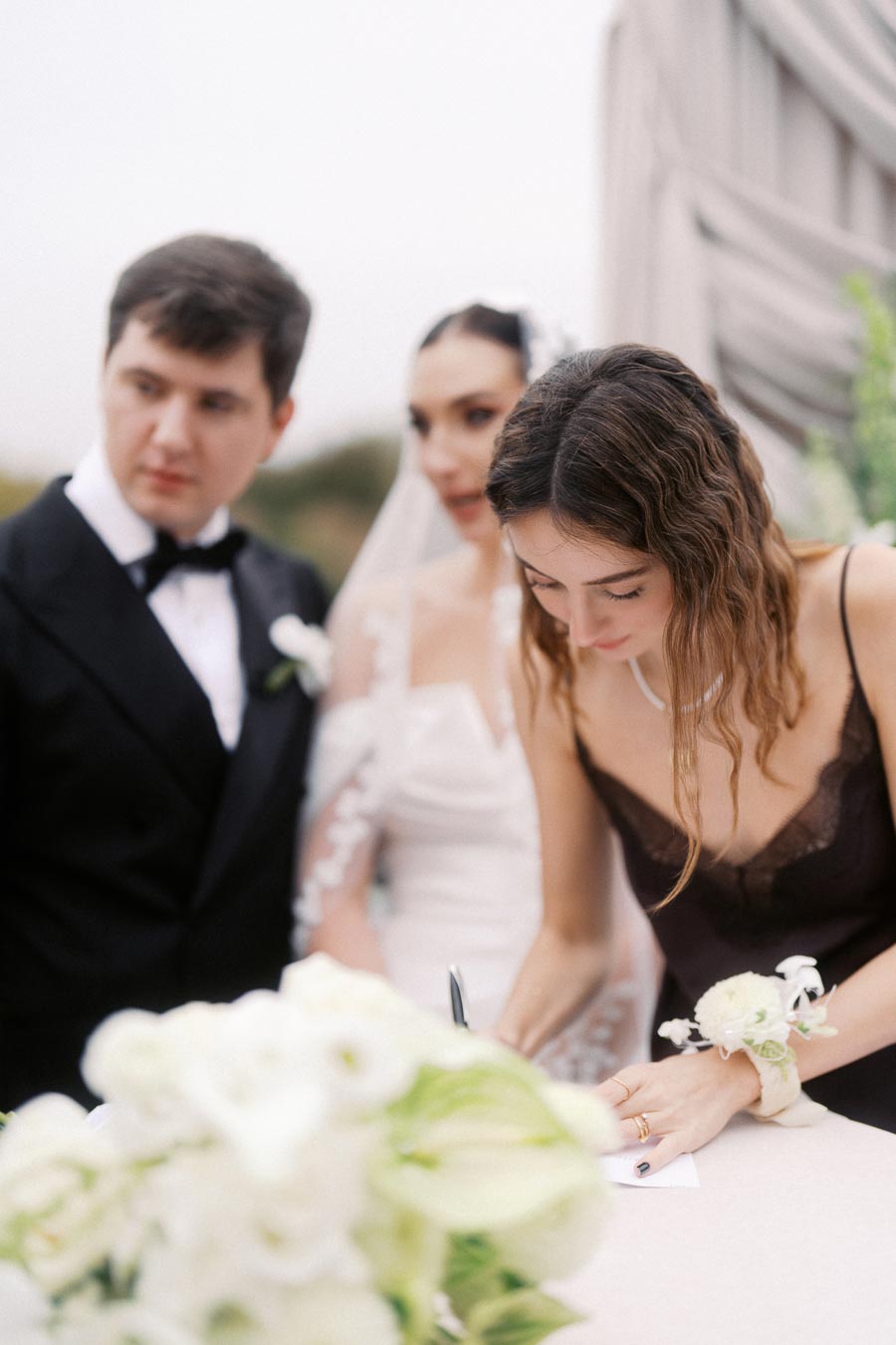 A woman in a bridesmaid dress signs a document at an outdoor wedding ceremony, with a bride in a white gown and groom in a tuxedo looking on, surrounded by white floral arrangements.