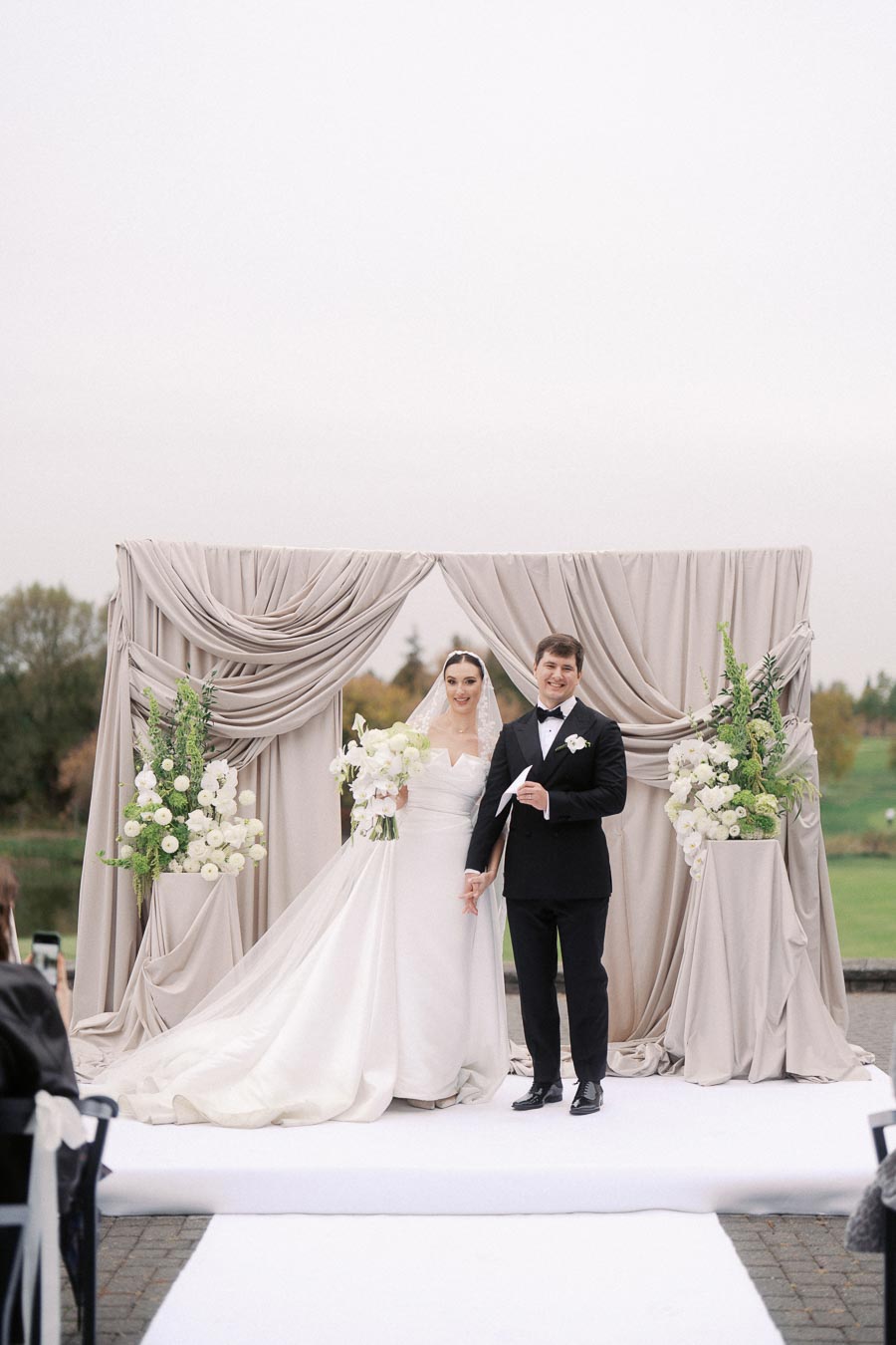 Elegant outdoor wedding ceremony with a bride in a white gown and veil, and a groom in a black tuxedo, standing in front of a draped floral backdrop.