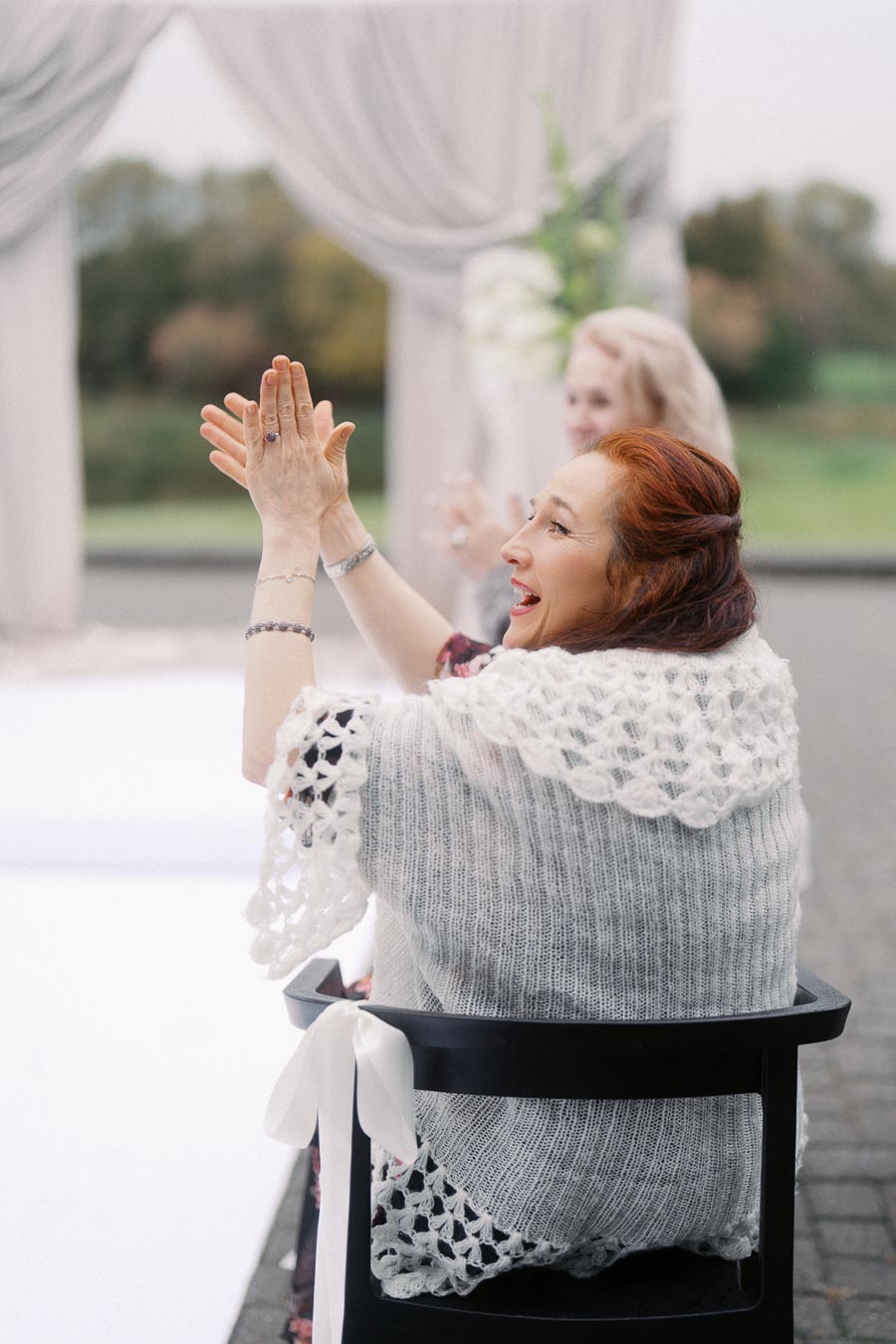 A woman with red hair and a white crocheted shawl clapping her hands joyfully at an outdoor event with draped fabric and greenery in the background.