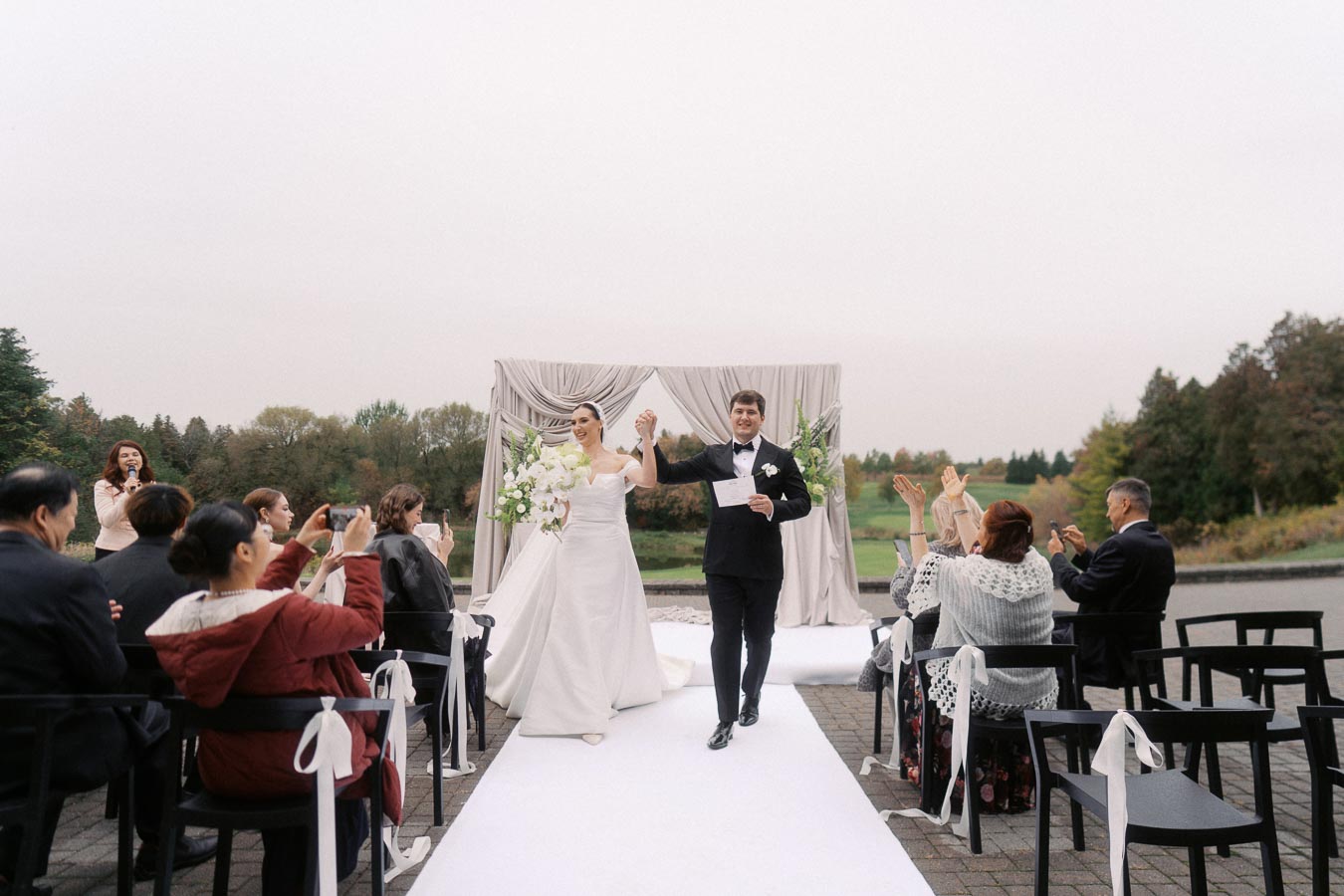 Outdoor wedding ceremony with a joyful bride and groom walking down the aisle, surrounded by guests taking photos and celebrating the couple's special day.