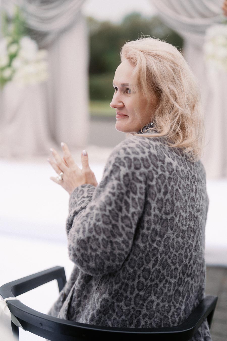 A woman with blonde hair sits on a black chair, wearing a leopard print jacket, looking to the side with a relaxed expression, against a blurred outdoor background.