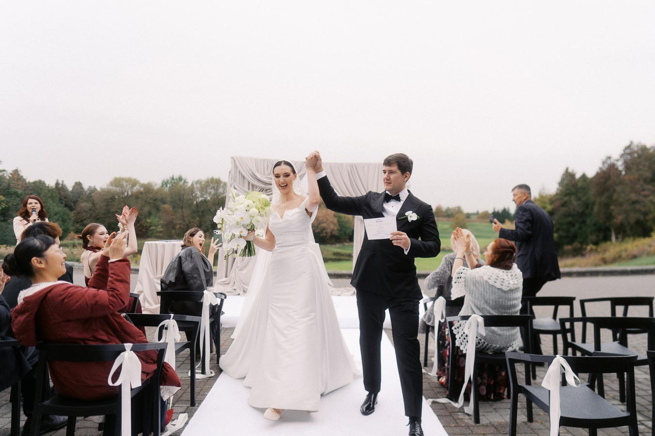 Bride and groom celebrating with guests at outdoor wedding ceremony, dressed in elegant attire, walking down an aisle adorned with white decorations, set against a scenic backdrop.