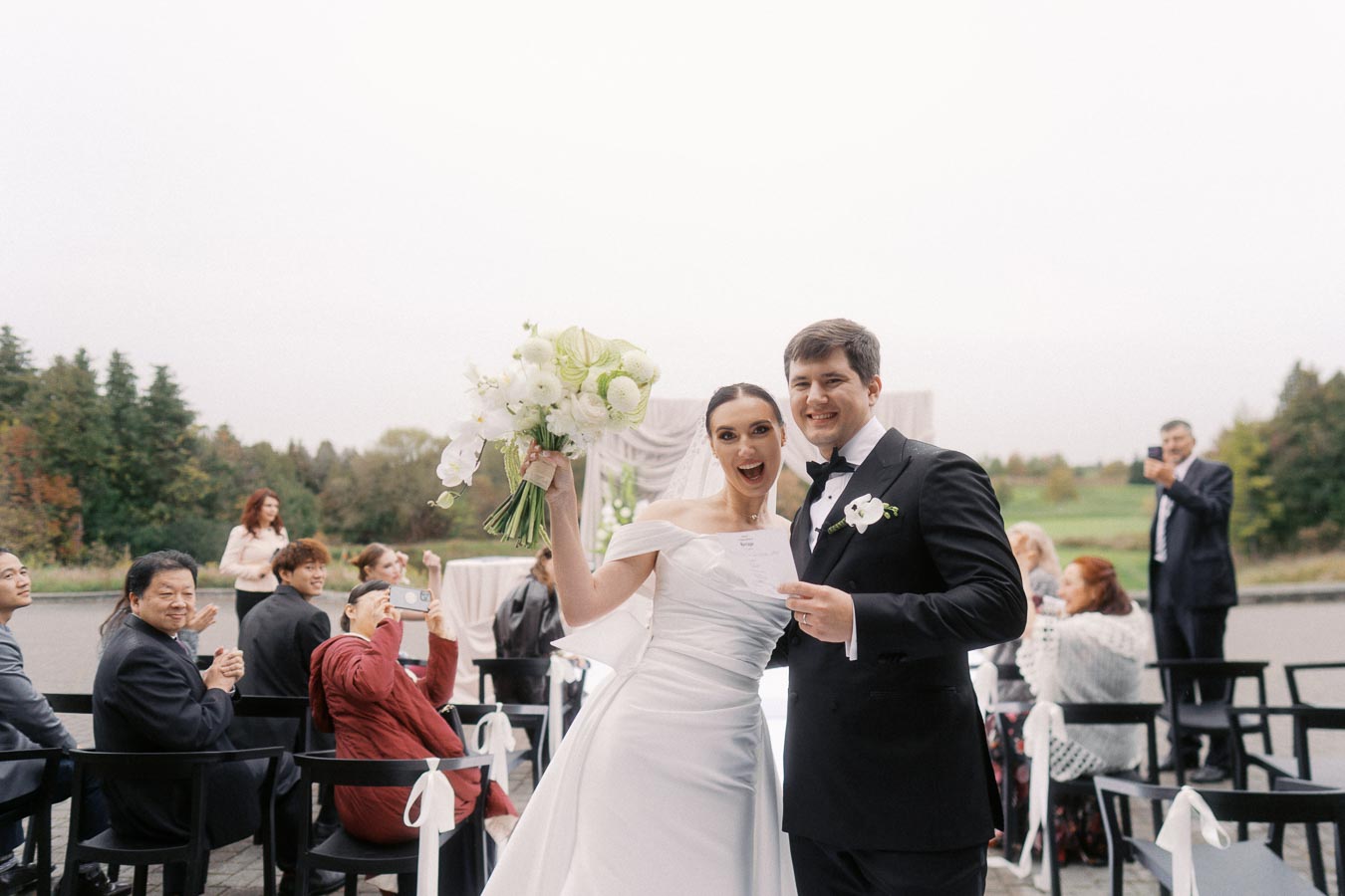 A joyful bride and groom celebrate their wedding ceremony outdoors, surrounded by smiling guests in formal attire. The bride holds a bouquet of white flowers, and the groom is in a classic black tuxedo. The setting features a scenic backdrop of greenery and a cloudy sky.