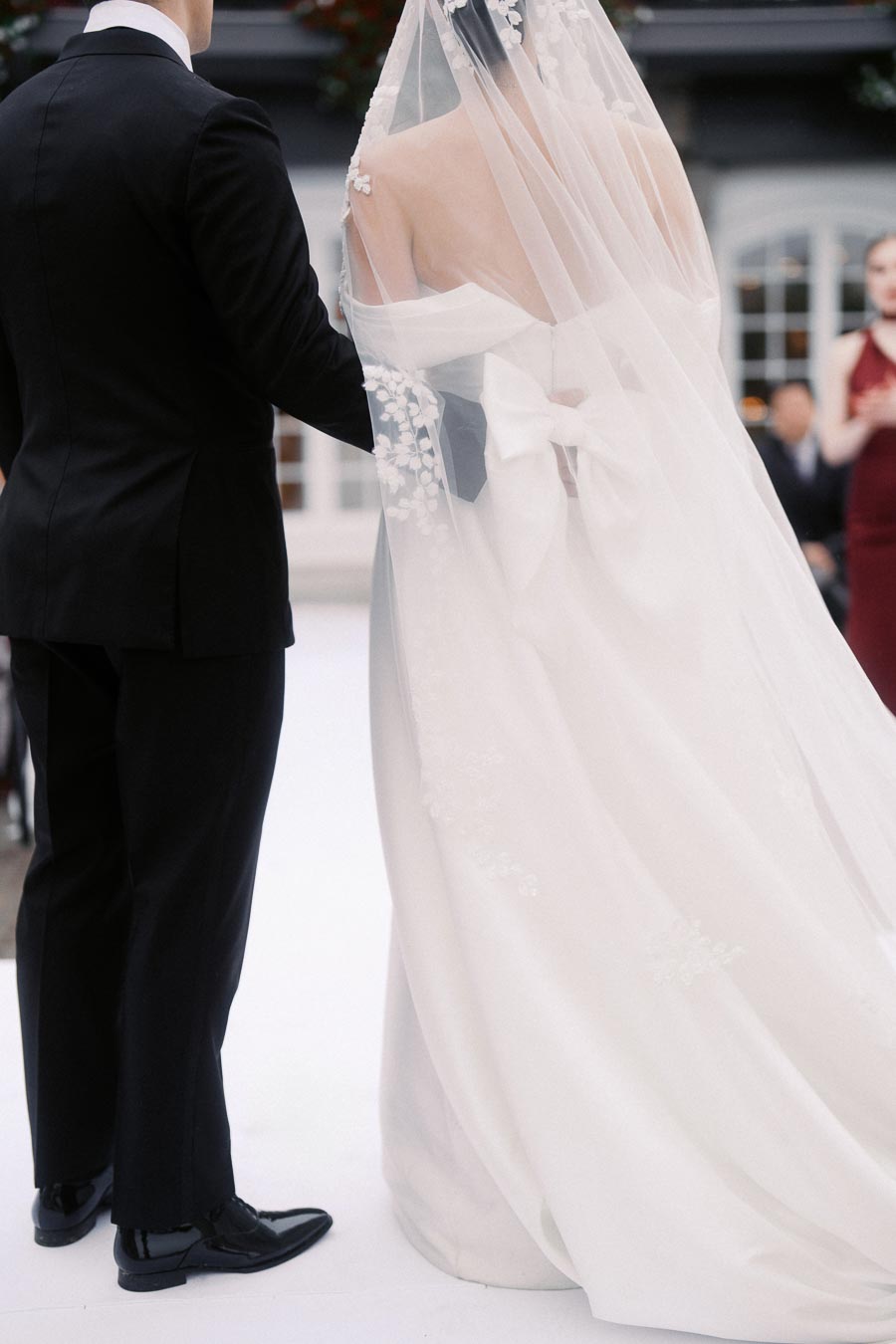 Elegant bridal couple standing together at a wedding ceremony, featuring the bride in a flowing white gown with a delicate lace veil, and the groom in a classic black tuxedo.
