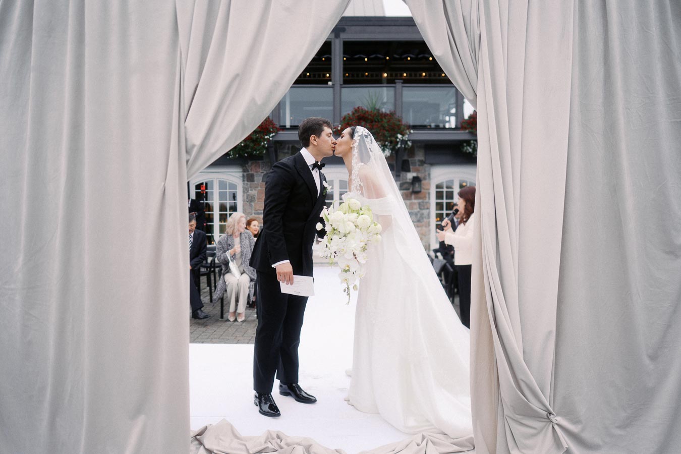 Elegant wedding ceremony scene with a bride in a white gown and groom in a black tuxedo sharing a kiss, framed by draped curtains and set against a charming stone building backdrop.