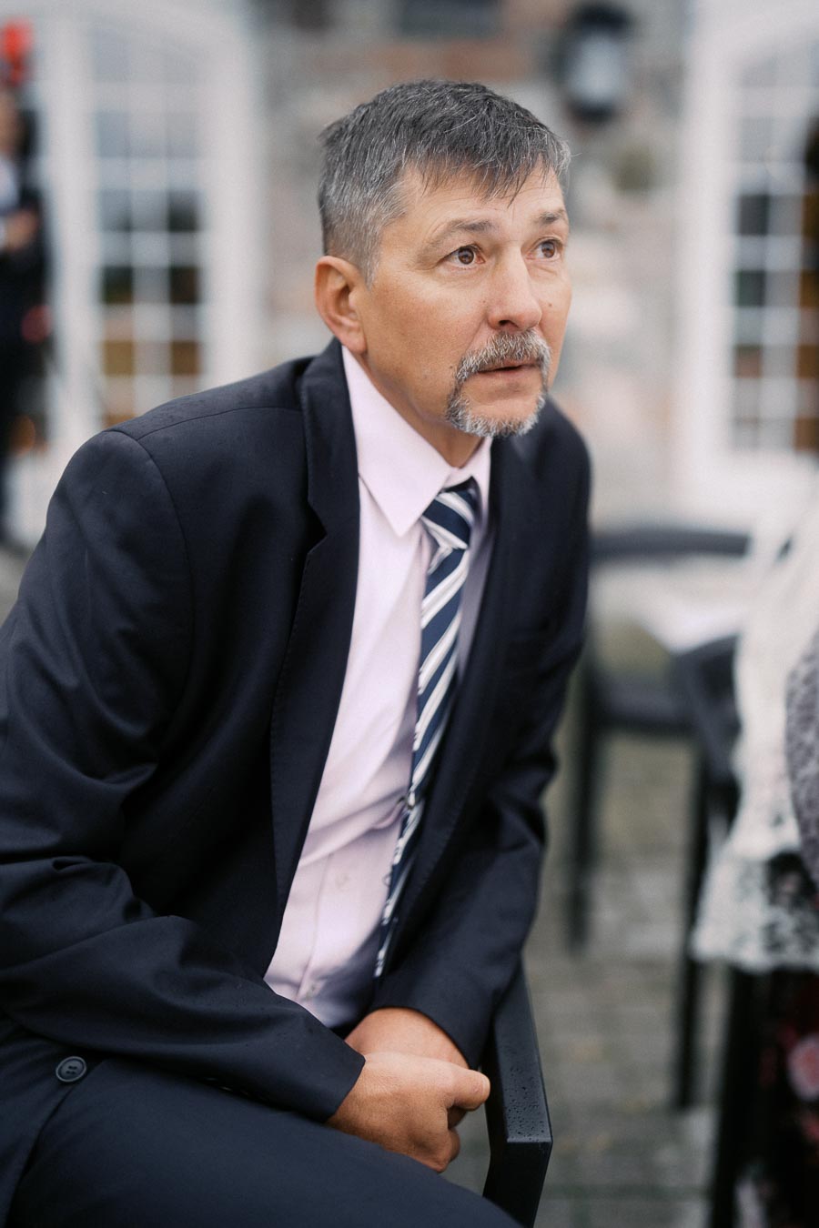 Adult man in a formal suit with a striped tie, sitting outside near a building with large windows, looking thoughtful.