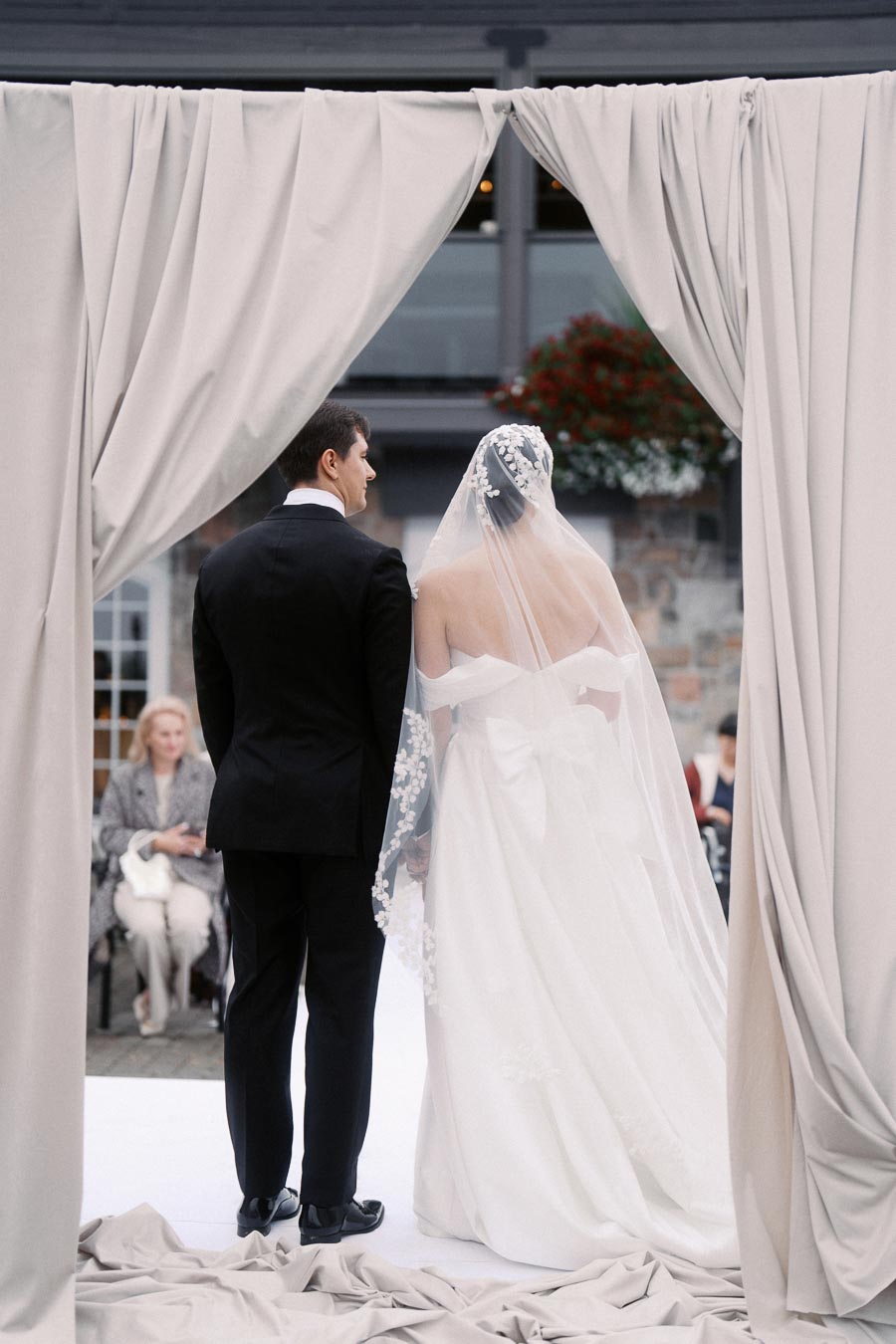 Bride and groom stand together under elegant draped curtains during an outdoor wedding ceremony, with guests seated in the background.