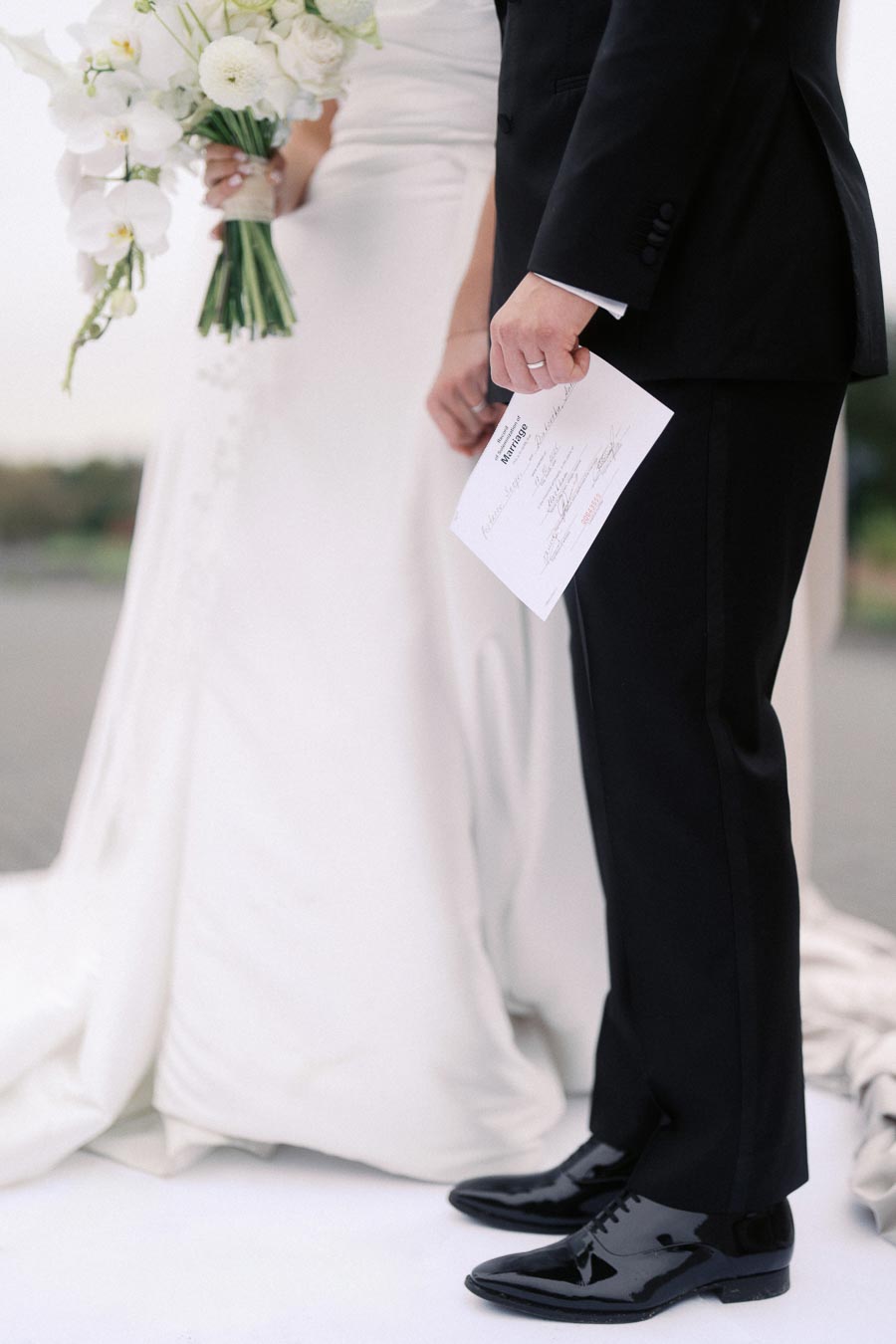 A newlywed couple stands side by side, the groom in a black suit holding a marriage certificate, while the bride in a white gown carries a bouquet of white flowers, capturing a moment of their wedding day celebration.