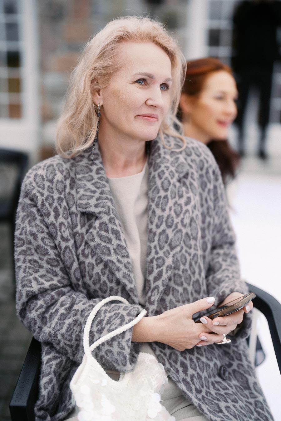 Middle-aged woman in a gray leopard-print coat holding a smartphone, sitting in an outdoor setting with a blurred background.