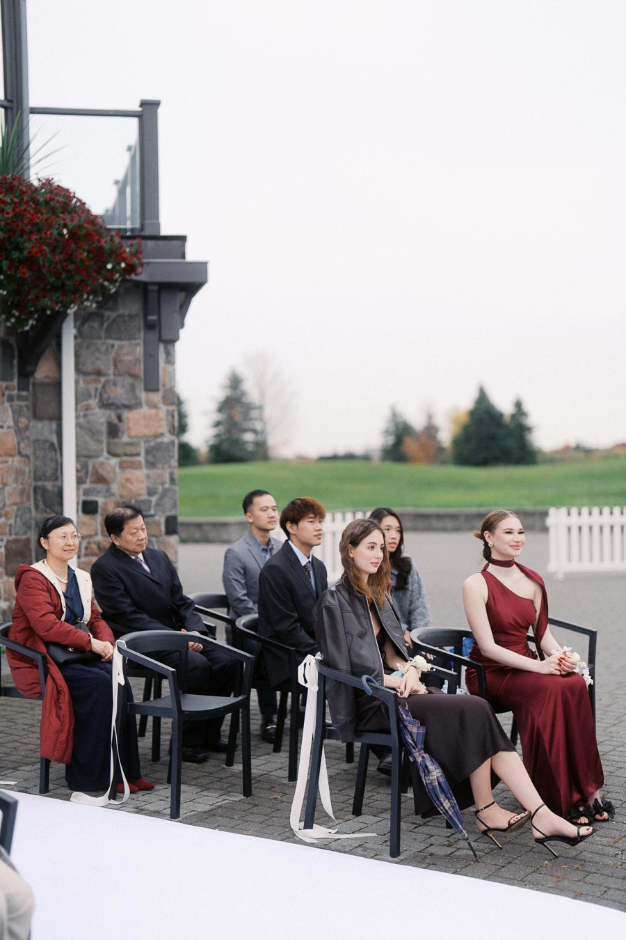 Guests seated outdoors at a formal event, wearing elegant attire. The setting includes a stone wall and flower arrangements, with a green landscape in the background.