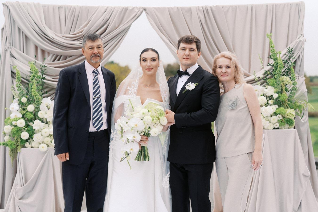 Wedding ceremony with bride and groom posing with family under elegant drapery, surrounded by white floral arrangements.