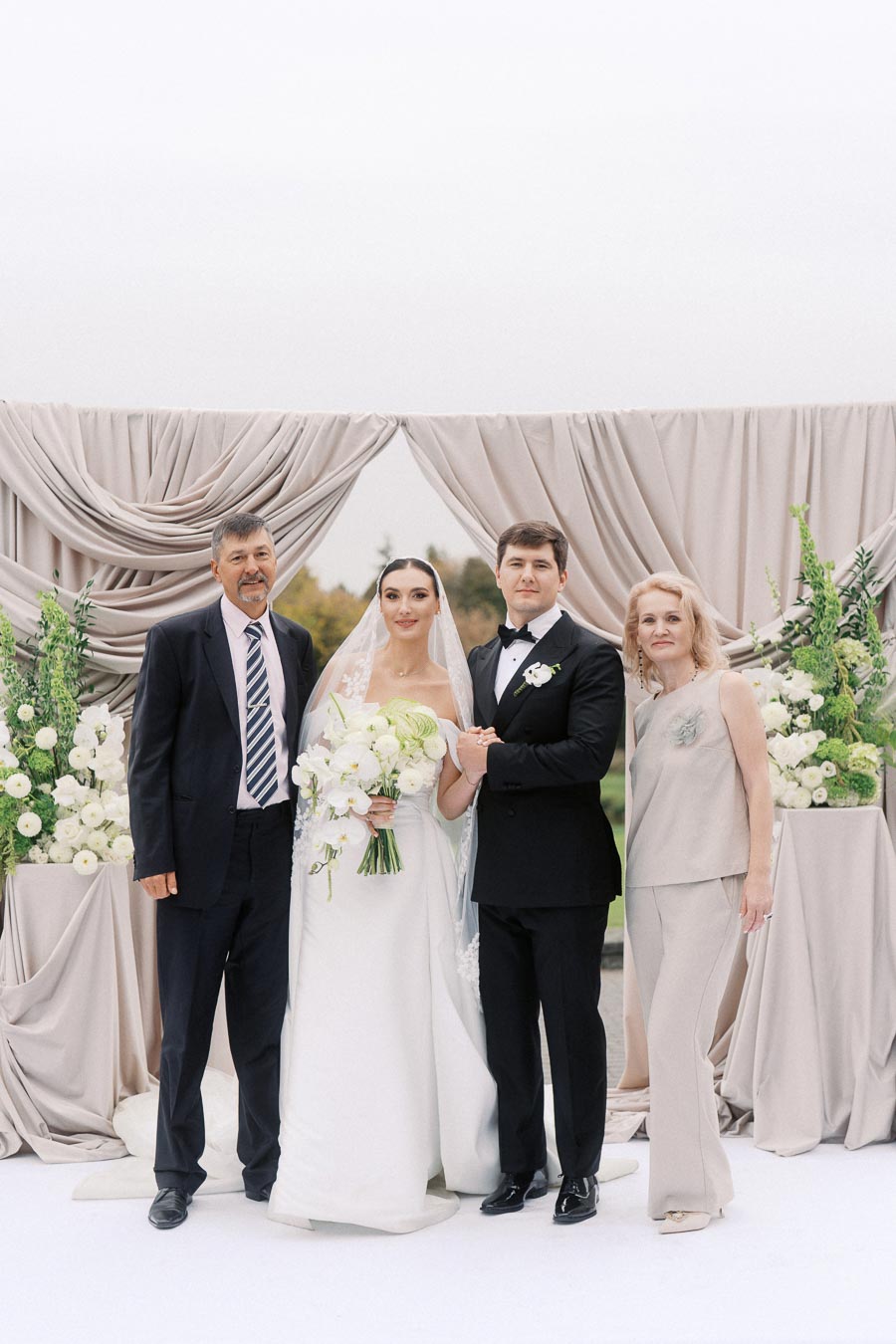 A bride and groom smiling with two guests at an outdoor wedding, standing in front of an elegant backdrop with draped curtains and floral arrangements.