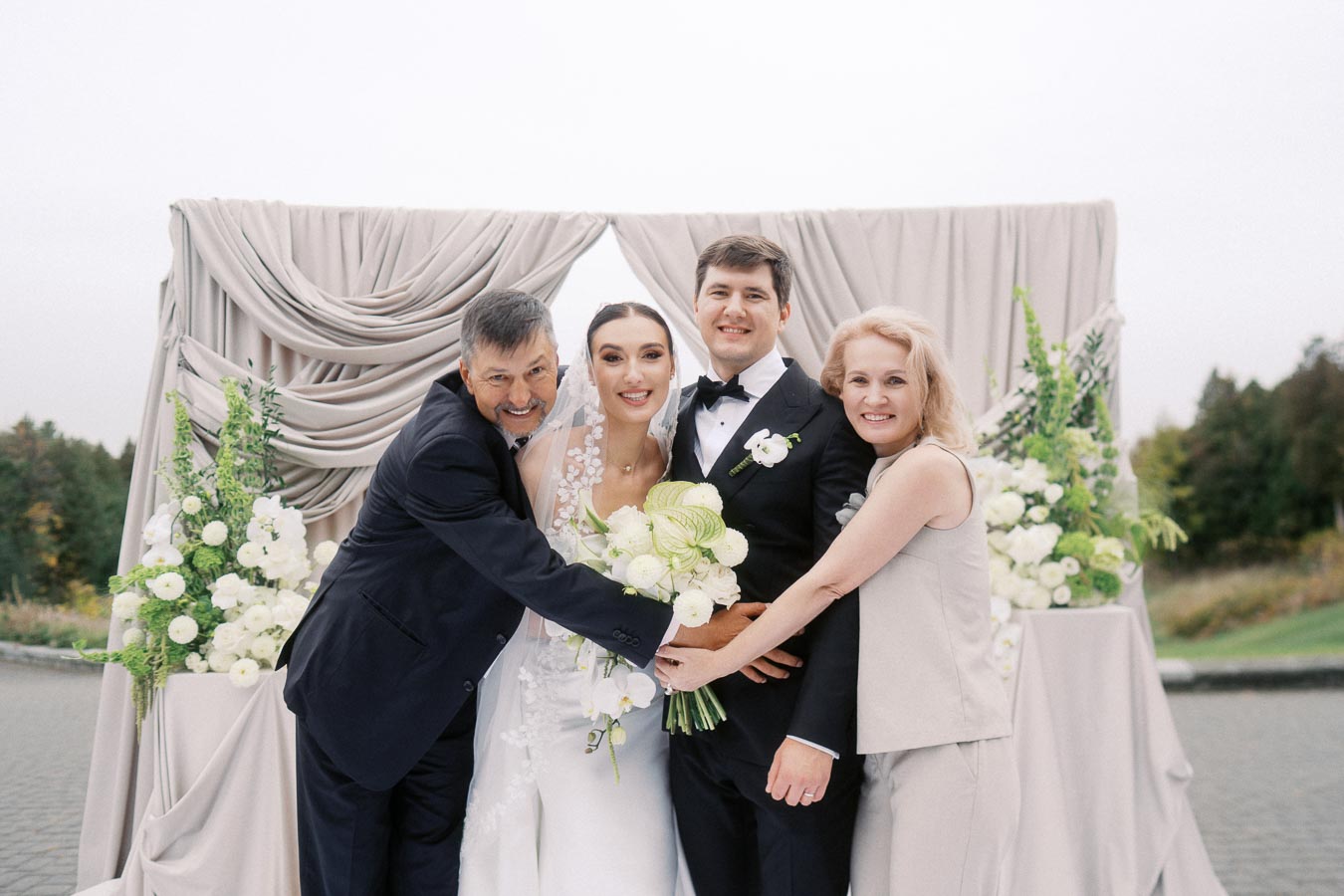 A joyful wedding photo featuring a bride and groom standing with two guests, posing happily in front of an elegant floral arch. The bride is wearing a beautiful white gown and veil, holding a bouquet of white flowers, while the groom is in a black tuxedo. The setting is outdoors with greenery in the background.