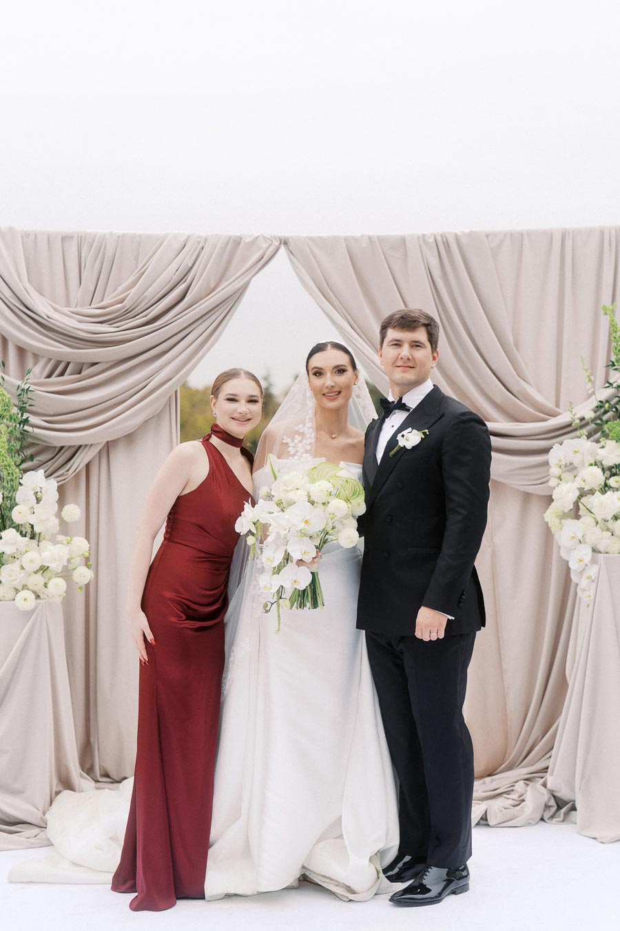 Wedding couple stands with bridesmaid in front of elegant draped backdrop. The bride holds a white floral bouquet and wears a classic white gown with a veil, while the groom is in a black tuxedo. The bridesmaid wears a red dress, and white flowers with greenery adorn the scene.