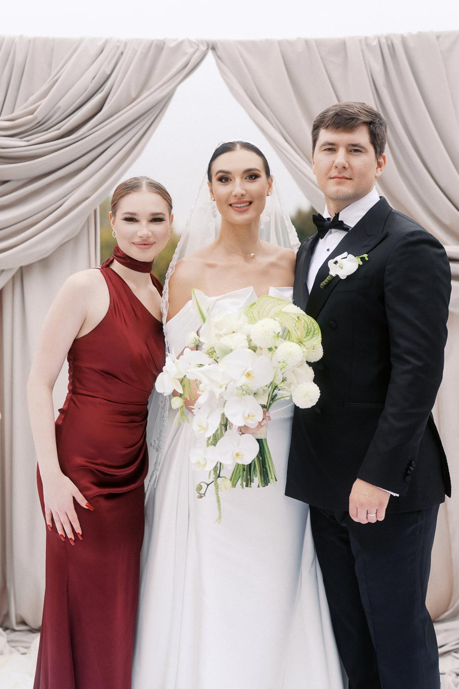 Wedding portrait featuring the bride in a white gown holding a floral bouquet, accompanied by a groom in a black tuxedo, and a bridesmaid in a burgundy dress, standing in front of elegant draped curtains.