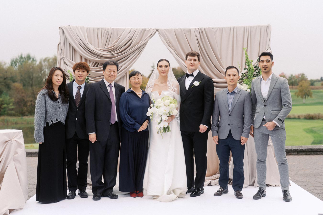 A diverse group of people in formal attire, posing together for a wedding photo in front of elegant drapery, with a scenic outdoor backdrop.