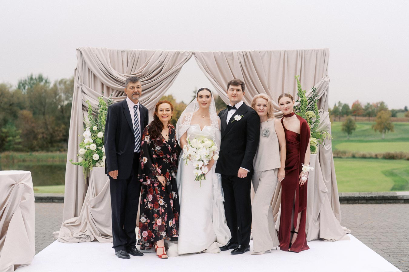 Wedding party group photo with bride in white gown and groom in black suit, surrounded by family, standing in front of elegant draped backdrop with floral arrangements, outdoor setting with trees and greenery.