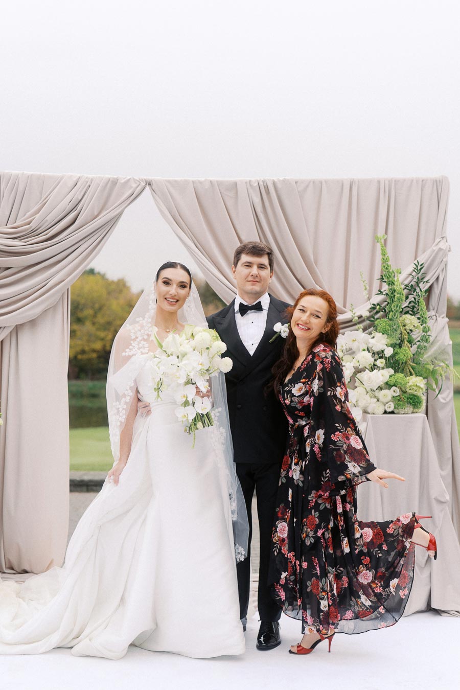 Bride and groom posing with guest during outdoor wedding ceremony, featuring elegant attire and floral decorations.