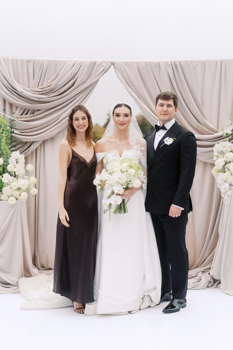 A bride and groom posing with a bridesmaid at a wedding ceremony, standing in front of elegant draped curtains and floral decorations. The bride is holding a bouquet of white flowers.