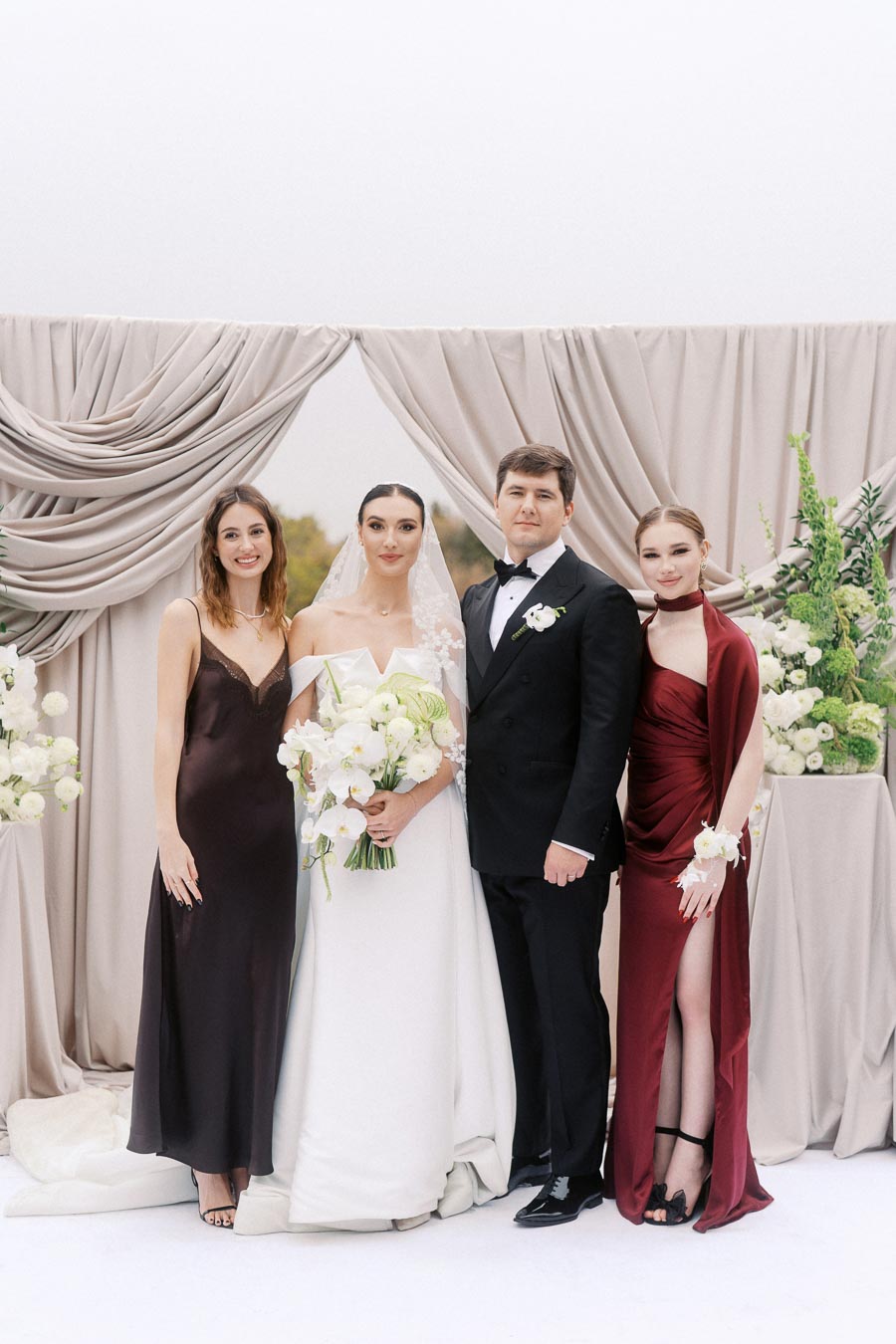 Wedding photo featuring a bride in an off-the-shoulder gown holding a bouquet, a groom in a tuxedo, and two bridesmaids in elegant dresses, standing in front of a draped fabric backdrop with floral arrangements.