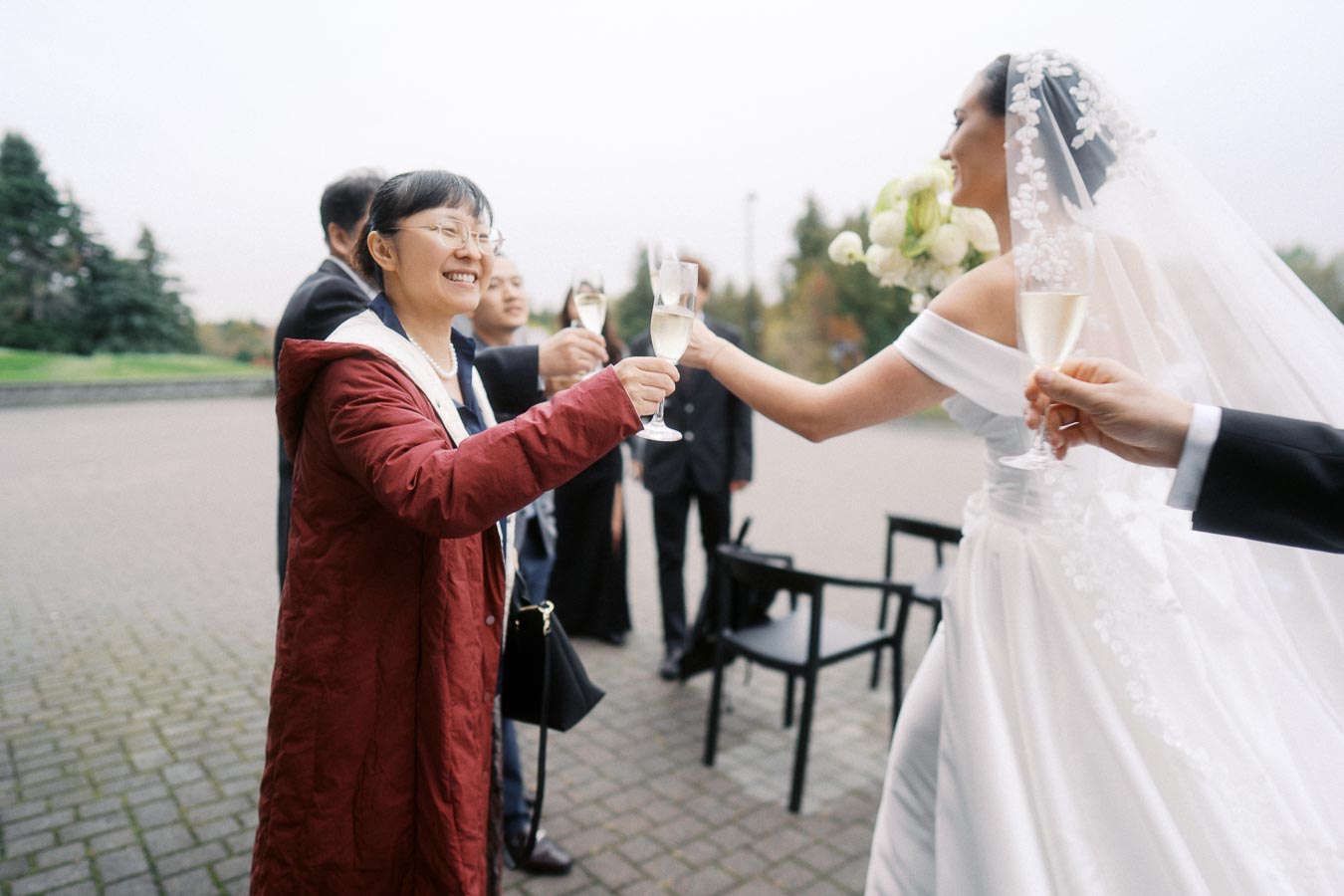 A bride in an elegant white dress with a lace veil toasts with guests outdoors, capturing a joyful moment at a wedding celebration.