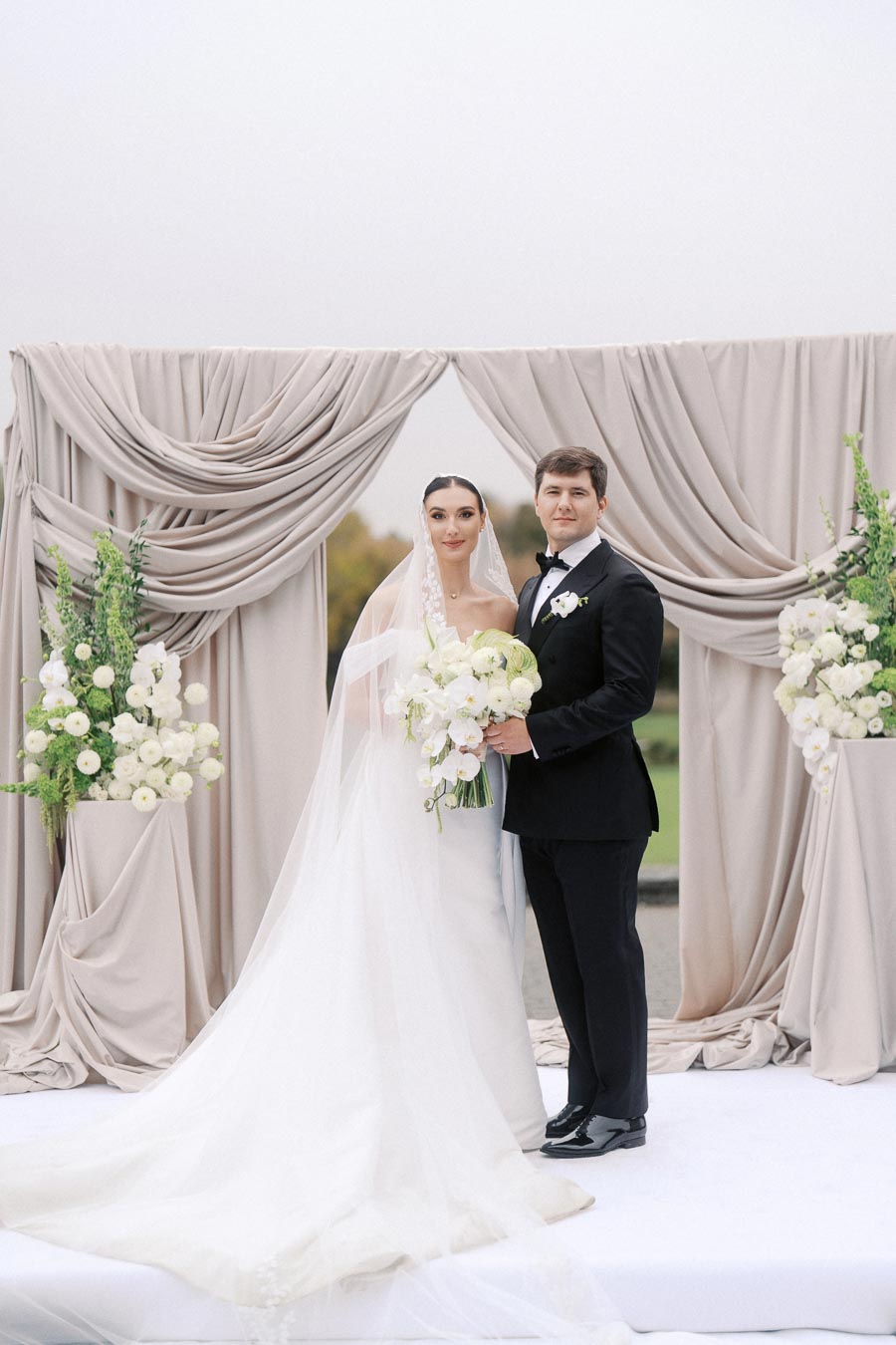 Elegant wedding couple posing in front of a beautifully draped backdrop with floral arrangements, showcasing a classic bridal gown and a stylish black tuxedo.