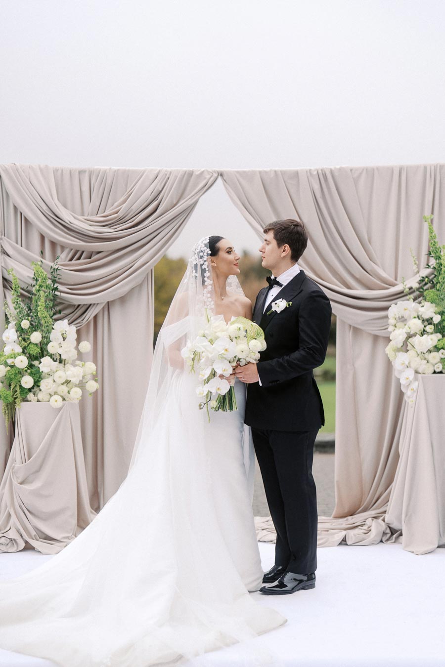 Elegant wedding couple standing together under a soft beige draped arch, holding a bouquet of white flowers, capturing a romantic moment on their special day.