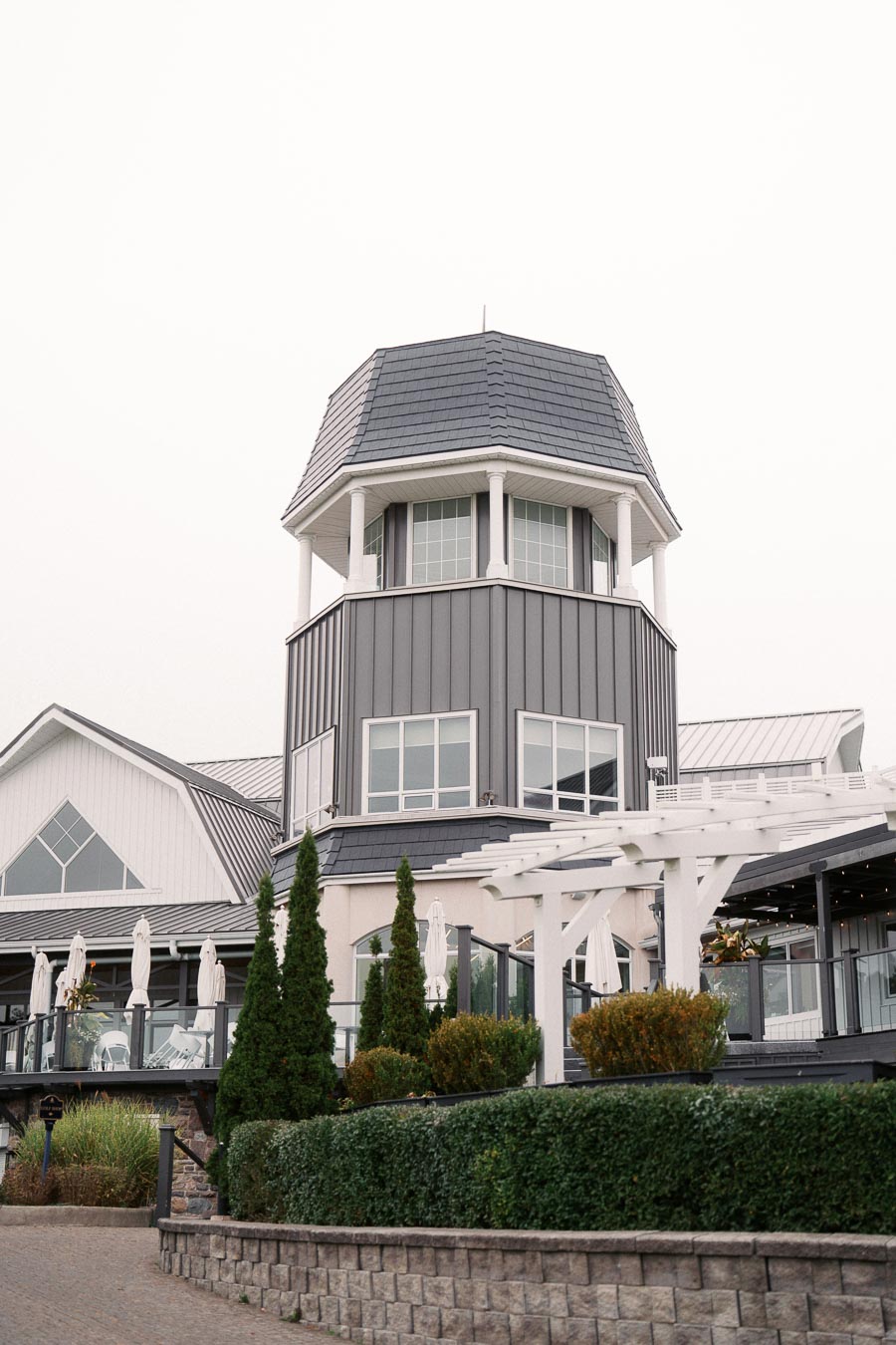 Modern tower building with a gray roof and large windows surrounded by greenery and a white pergola, highlighting contemporary architectural design.