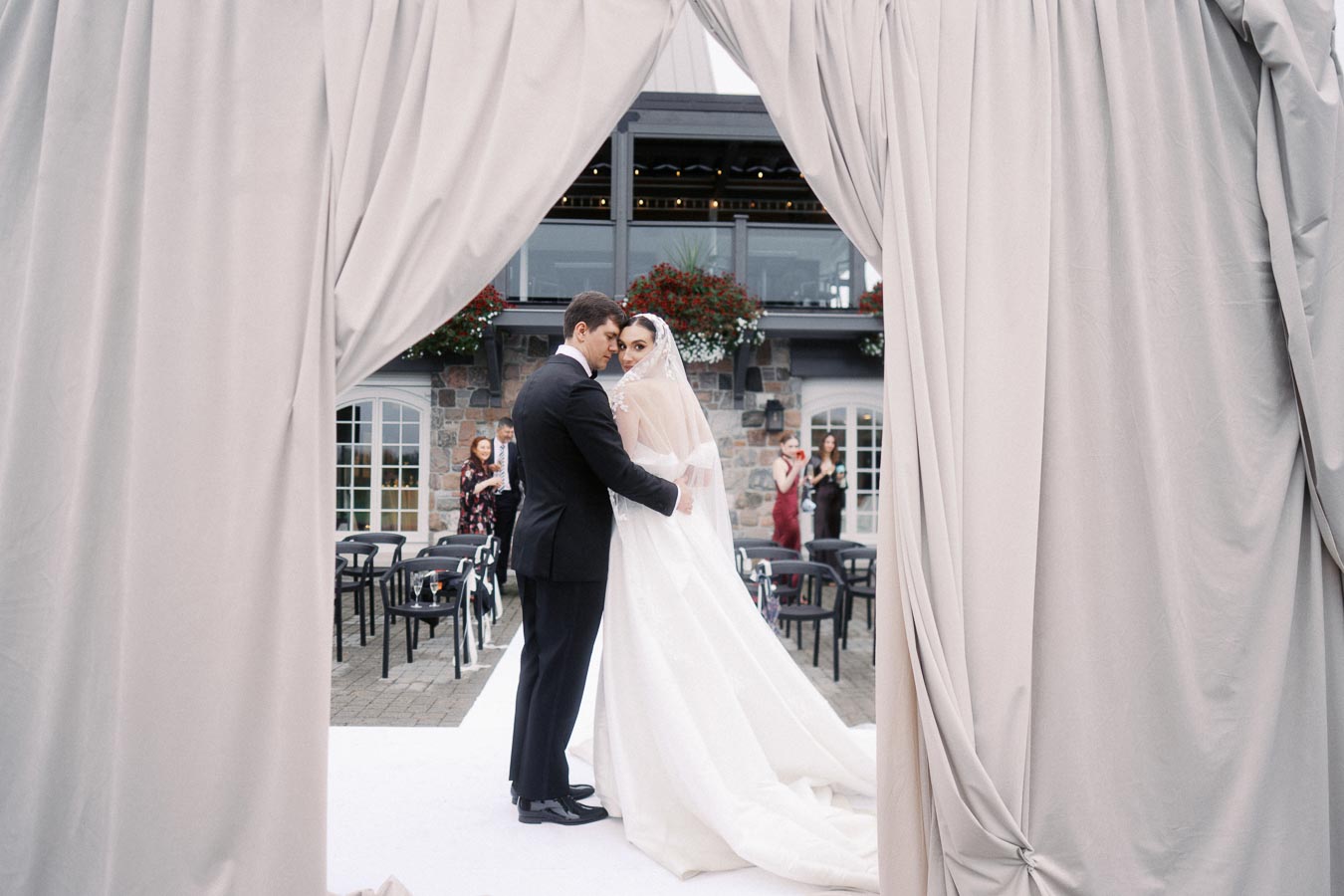 Elegant wedding couple posing in front of an outdoor venue with stone walls and draped curtains, capturing a romantic and intimate moment on their special day.