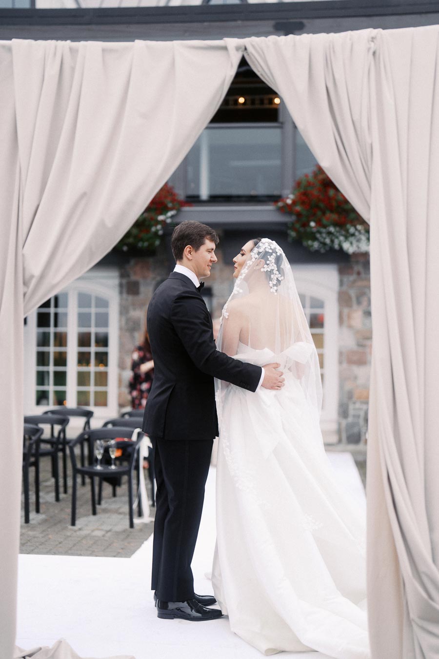 Bride and groom standing together under a wedding arch, wearing a floral veil and black suit, embracing during an outdoor ceremony.