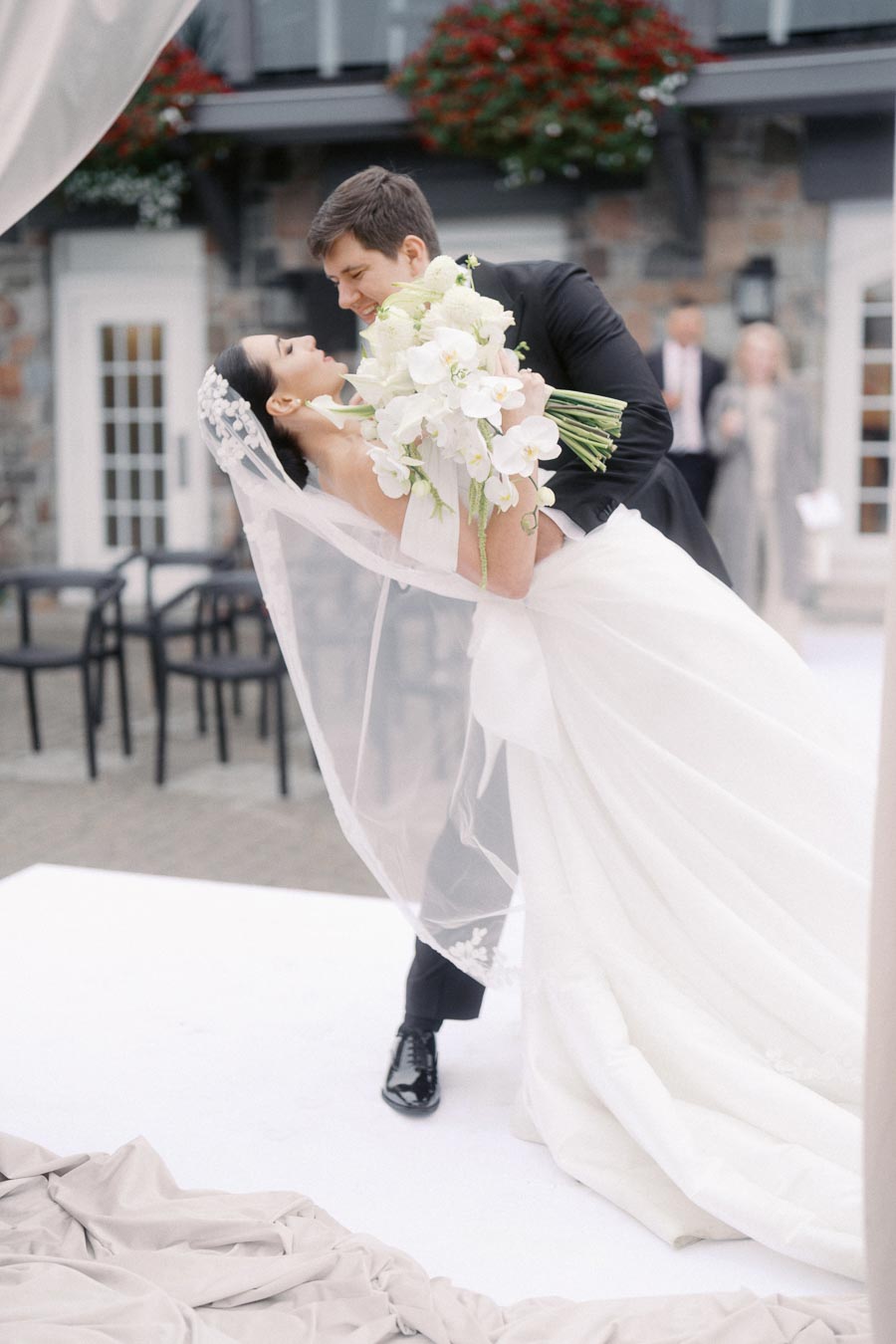 Romantic wedding moment with groom in black suit lovingly dipping bride in elegant white gown holding bouquet, set outdoors against charming stone building backdrop.